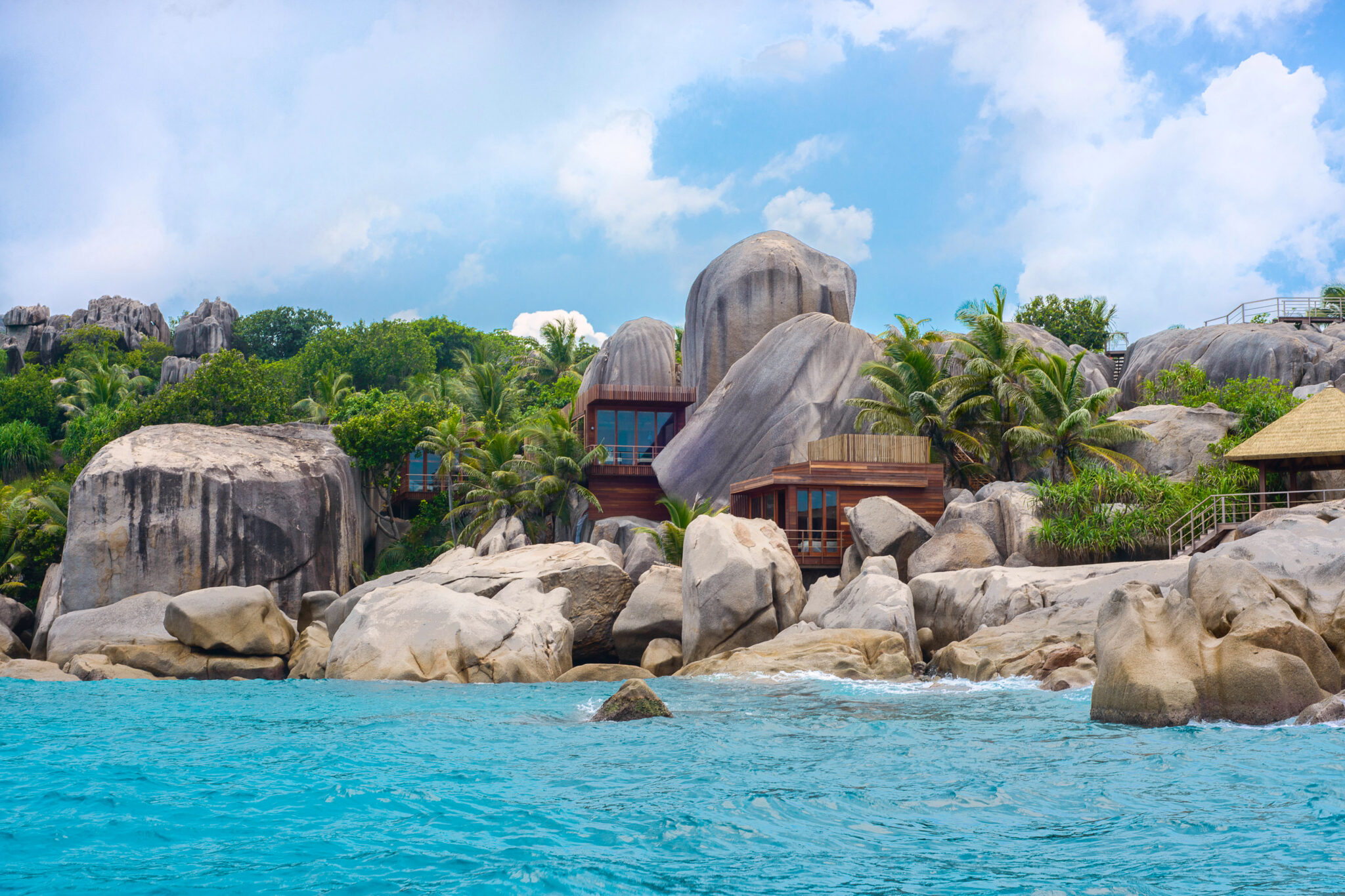 Granite boulders line a turquoise shore below a tropical spa pavilion, with palm trees and thatched roofs in sun.