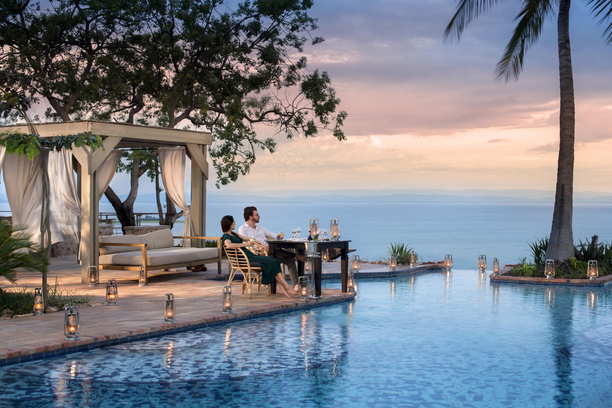 Guests sit beside an infinity pool at sunset, with a draped cabana, palm tree, and calm ocean horizon beyond.