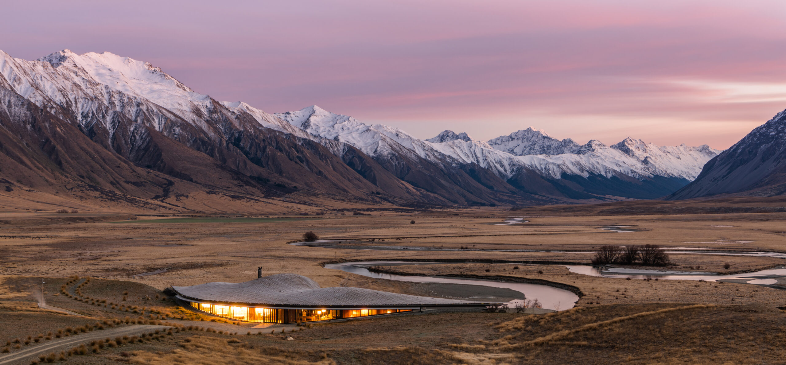 A low lodge glows in a broad valley as a winding road leads toward snowcapped mountains under a pink sky.