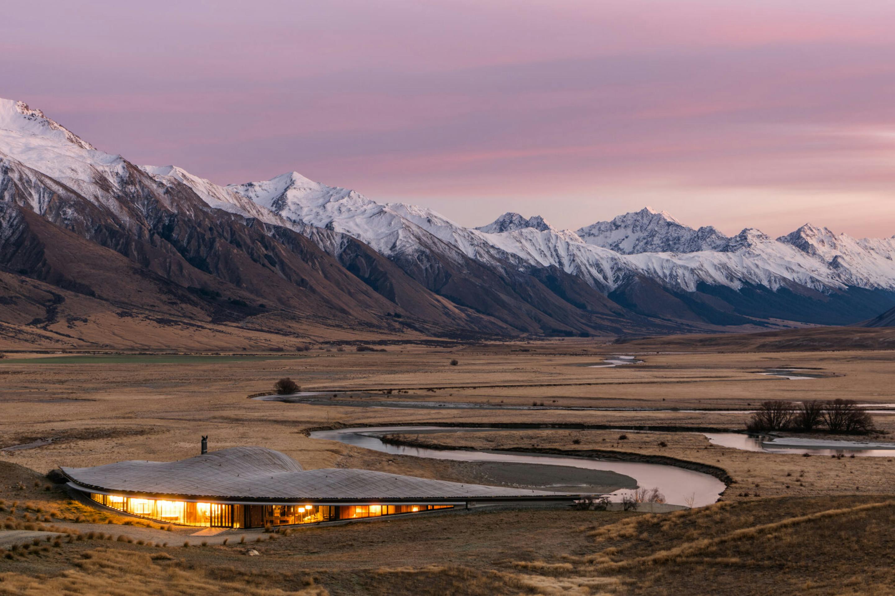 A low lodge glows in a broad valley as a winding road leads toward snowcapped mountains under a pink sky.