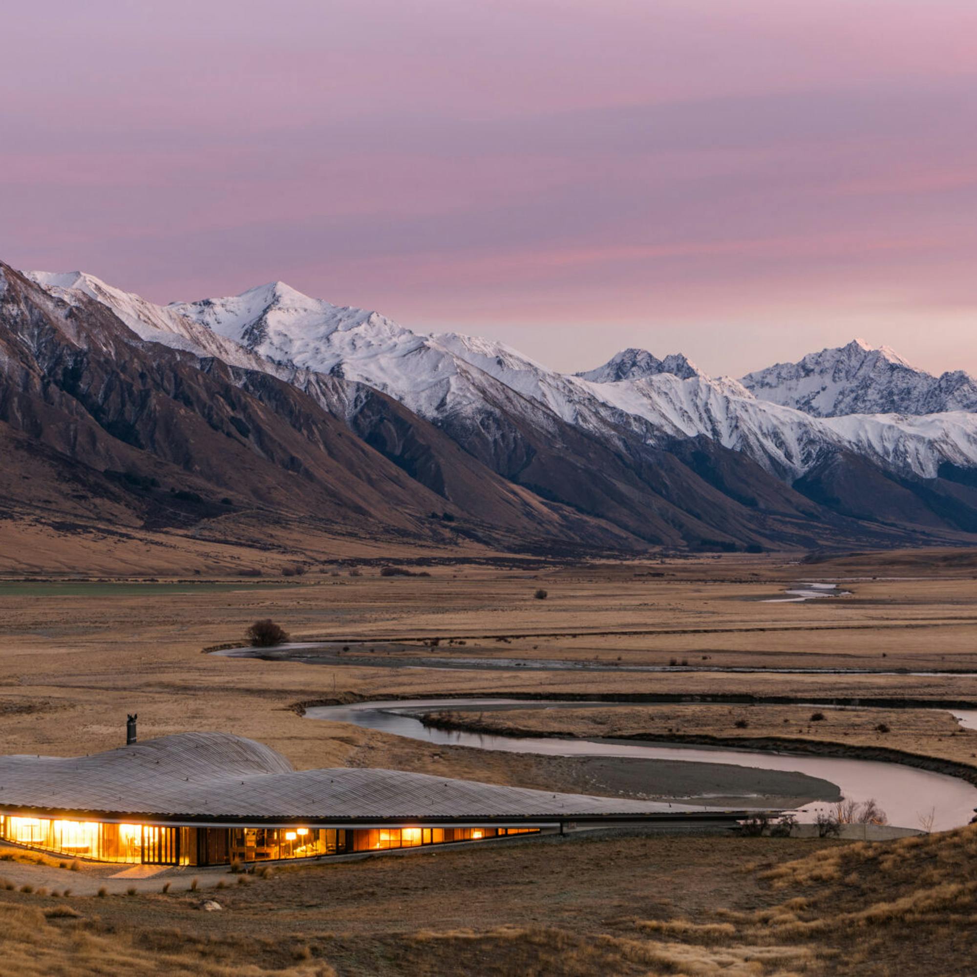 A low lodge glows in a broad valley as a winding road leads toward snowcapped mountains under a pink sky.