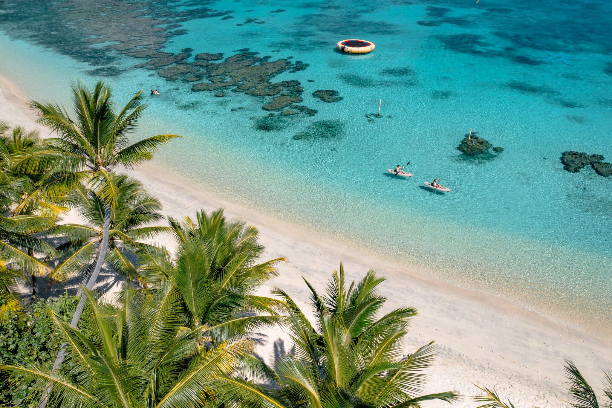 Aerial view of a palm-lined white-sand beach and turquoise lagoon dotted with small boats and coral patches.