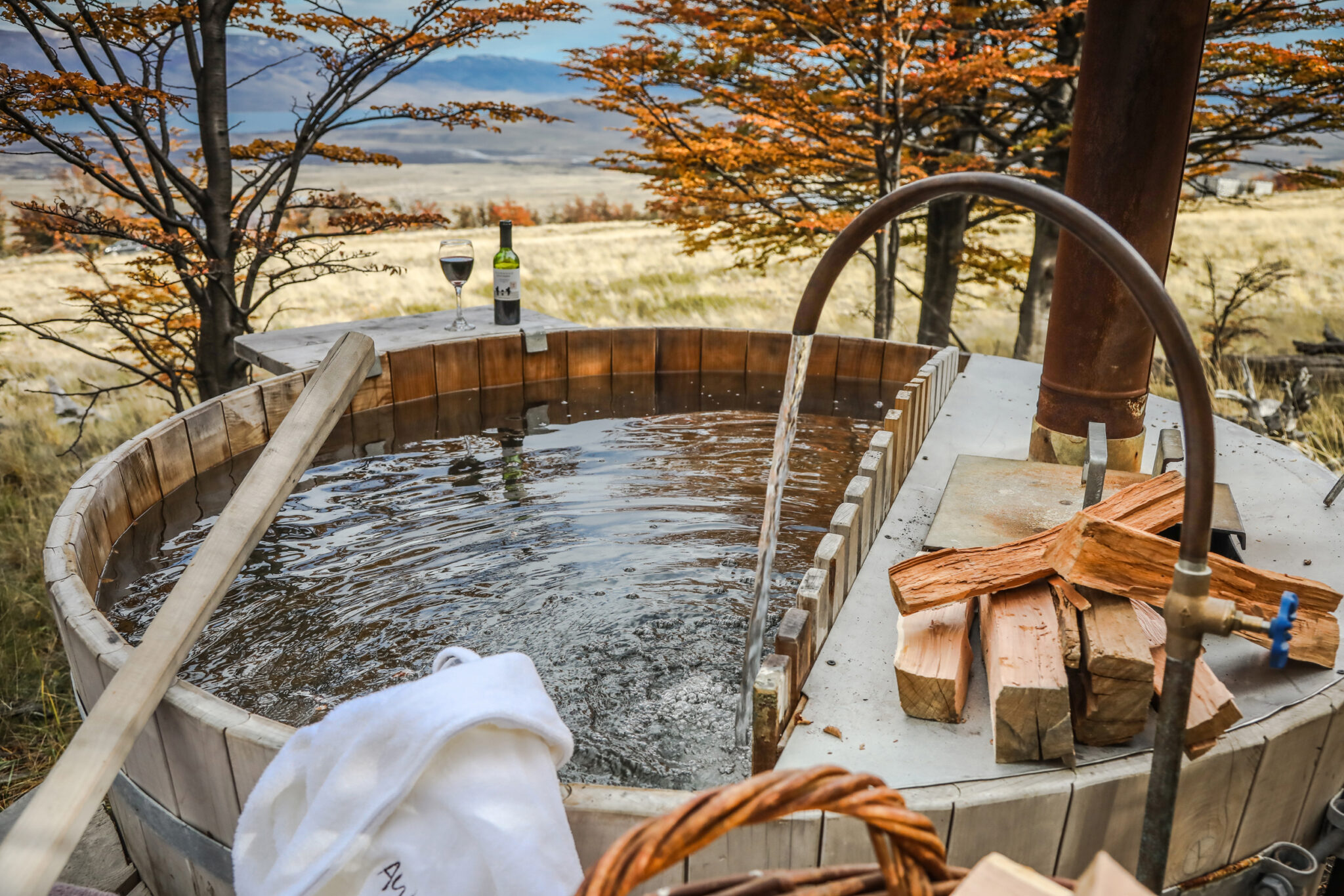 A wooden hot tub sits on a deck with towels and drinks, overlooking pale plains and golden trees in Patagonia.