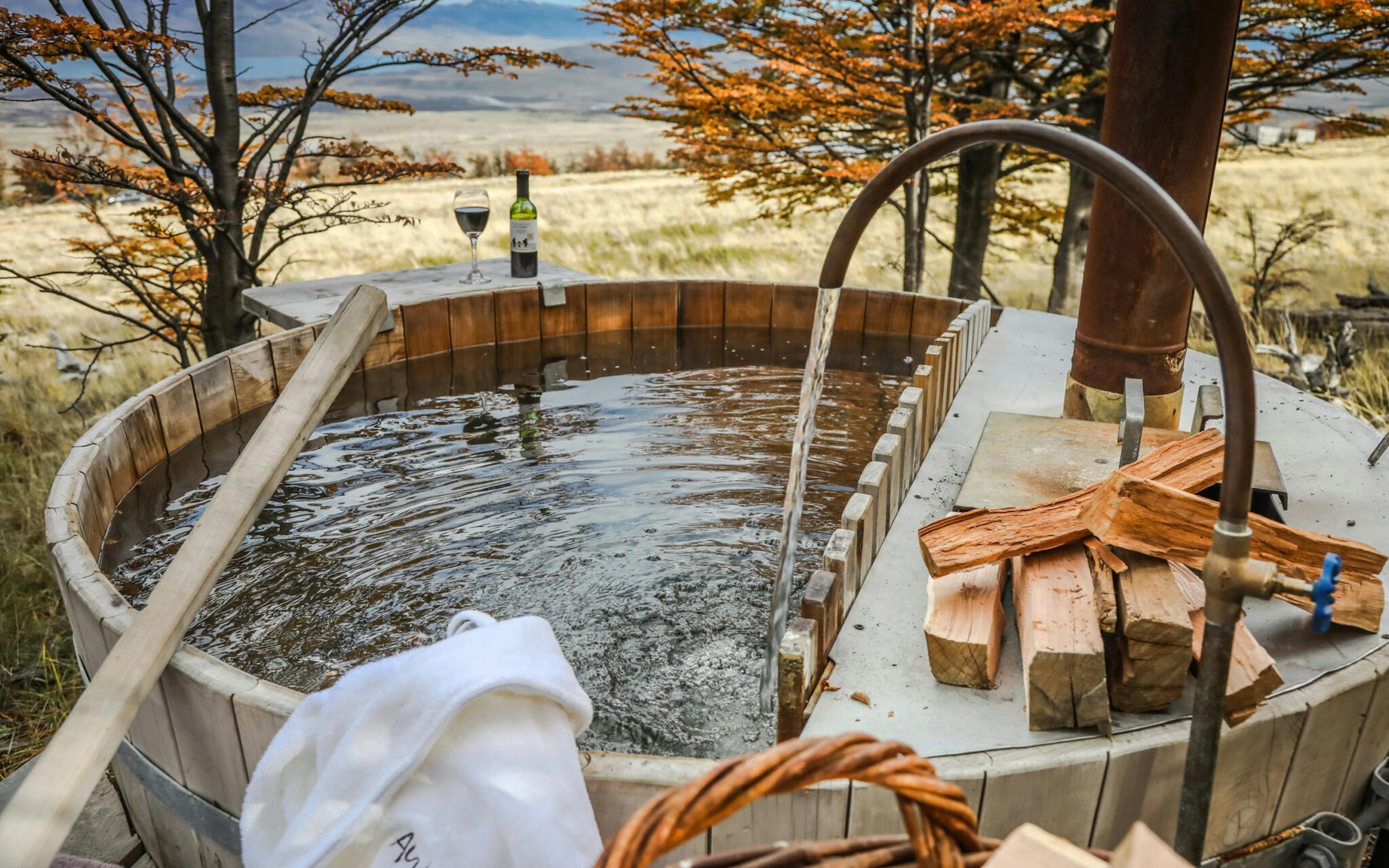 A wooden hot tub sits on a deck with towels and drinks, overlooking pale plains and golden trees in Patagonia.