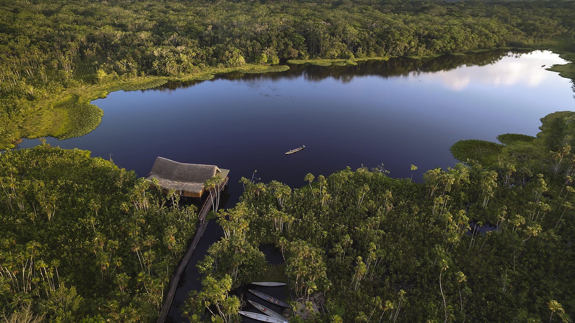 A thatched lodge sits on a wooden boardwalk at the edge of a dark rainforest lake with still water, framed by treetops.