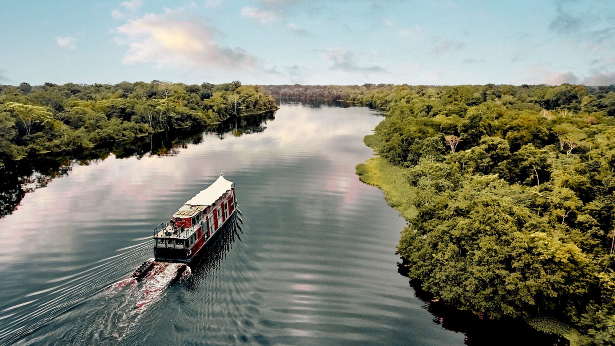 A river cruise ship leaves a rippled wake as it glides between lush green banks, reflected in smooth dark water.