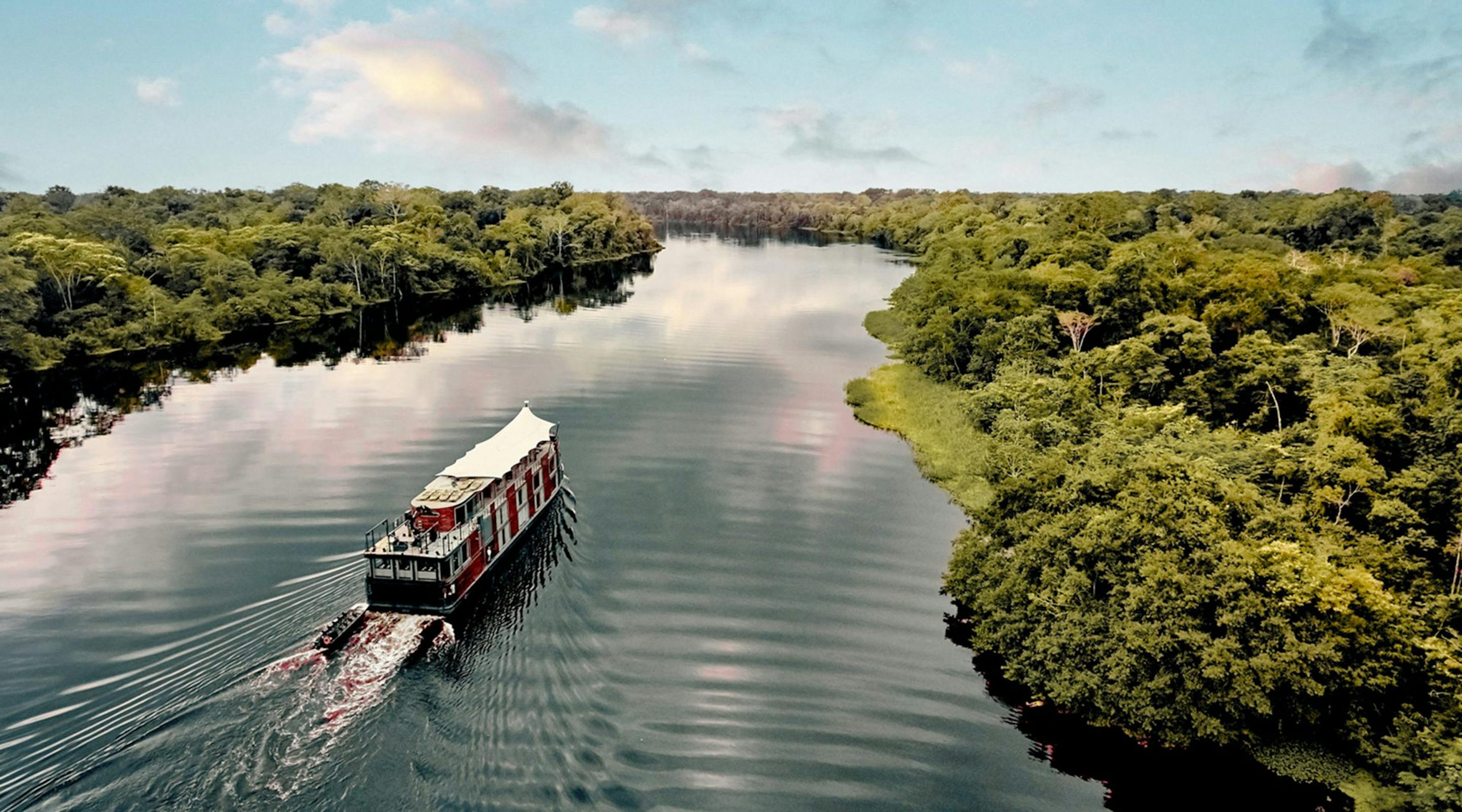 A river cruise ship leaves a rippled wake as it glides between lush green banks, reflected in smooth dark water.