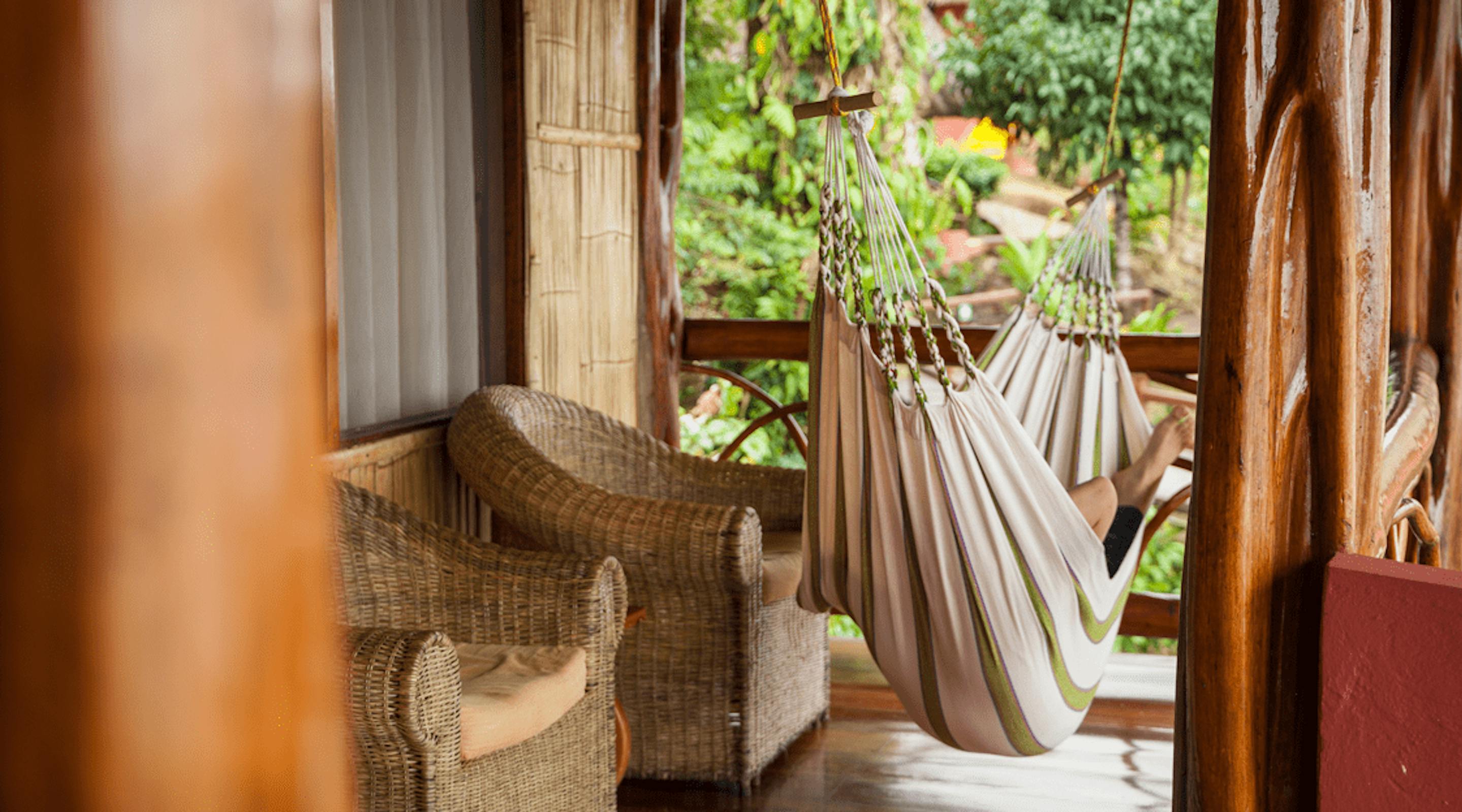 A striped hammock hangs on a wooden veranda beside wicker chairs, with rainforest greenery beyond in soft light.