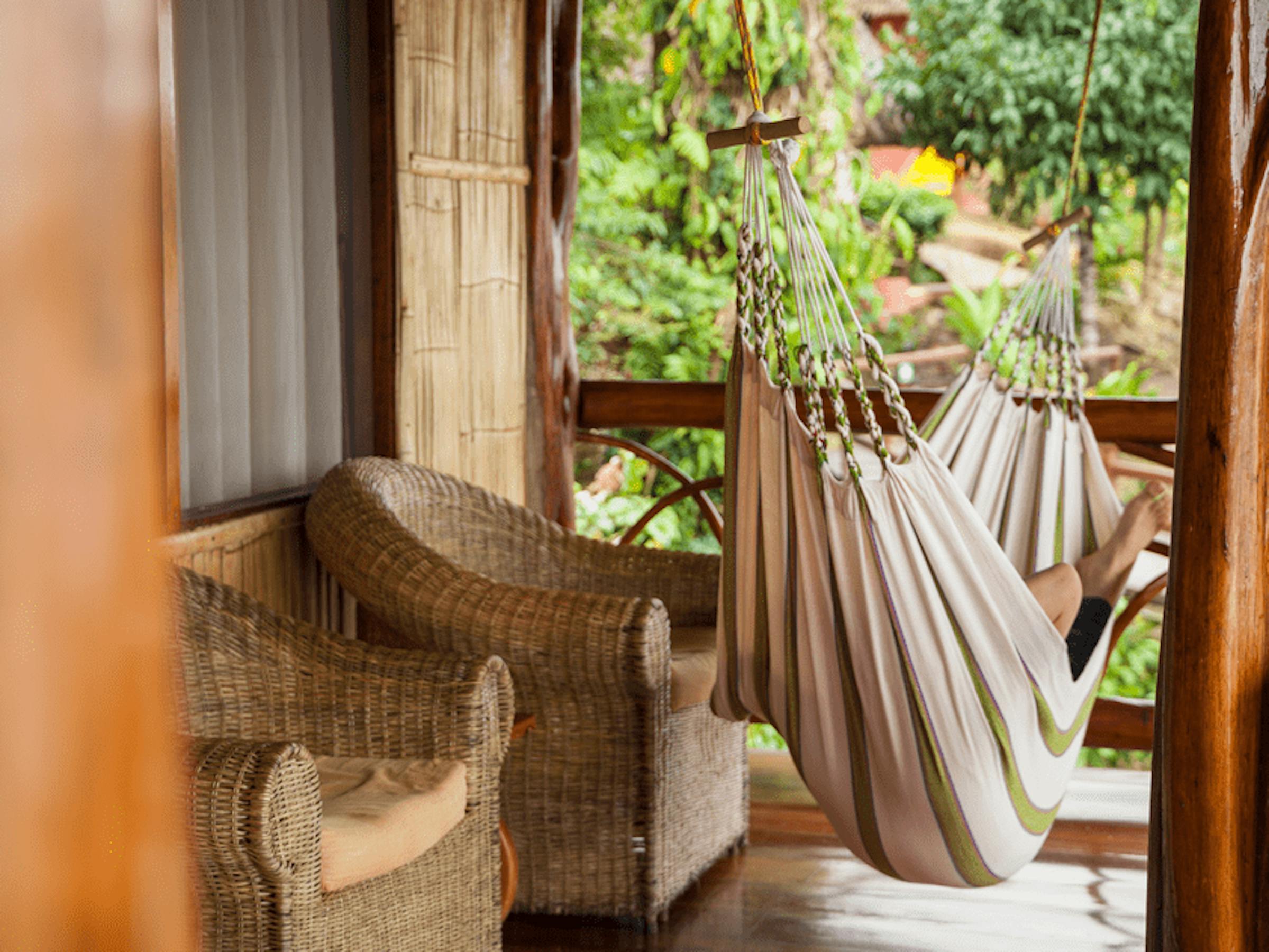 A striped hammock hangs on a wooden veranda beside wicker chairs, with rainforest greenery beyond in soft light.