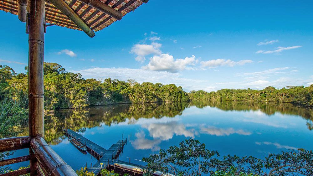 View from a wooden balcony over a mirror-still lake, with rainforest treeline and clouds reflected in blue water.