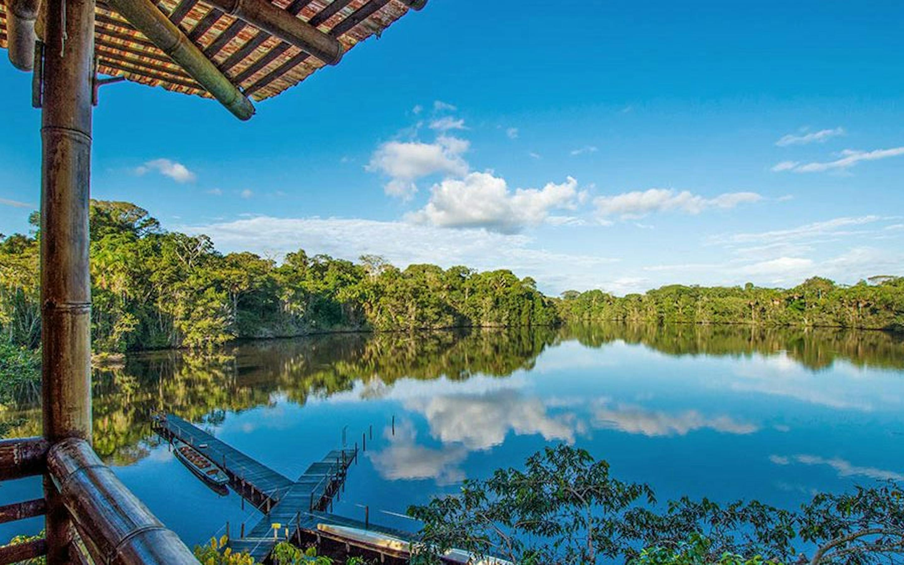 View from a wooden balcony over a mirror-still lake, with rainforest treeline and clouds reflected in blue water.