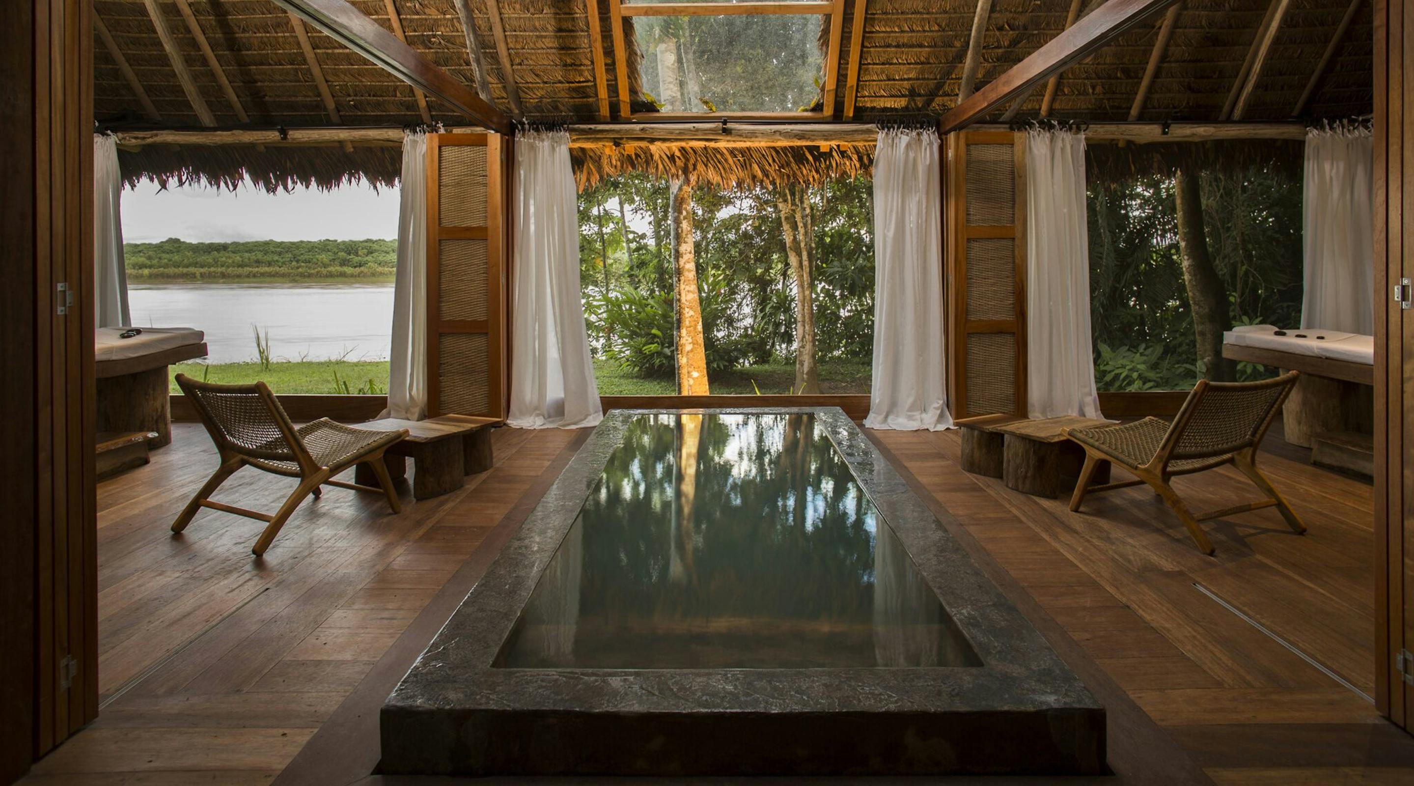 Indoor plunge pool sits between wooden loungers and open curtains, framing river water and jungle trees outside.