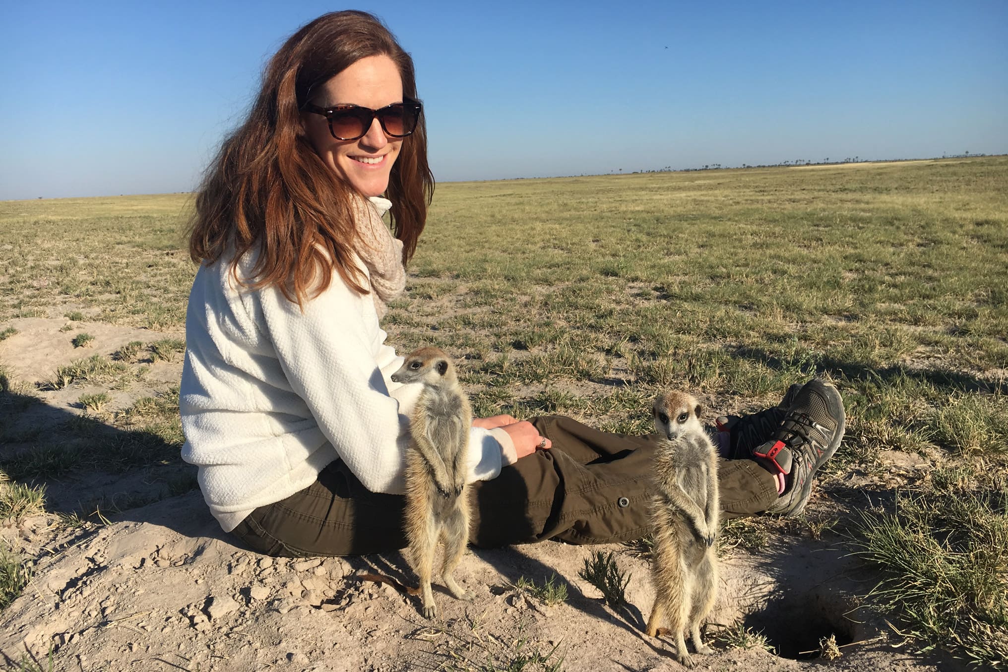 Traveler sits on sandy ground wearing sturdy hiking boots and sunglasses, watches meerkats next to her.