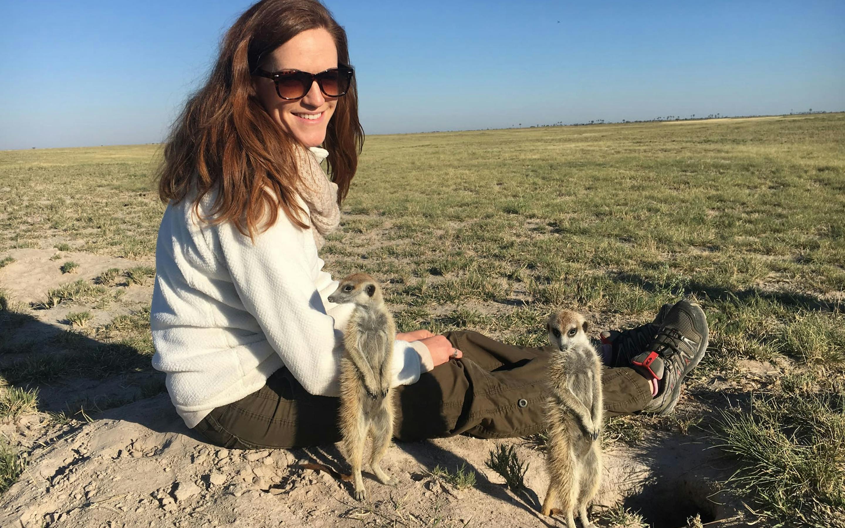 Traveler sits on sandy ground wearing sturdy hiking boots and sunglasses, watches meerkats next to her.