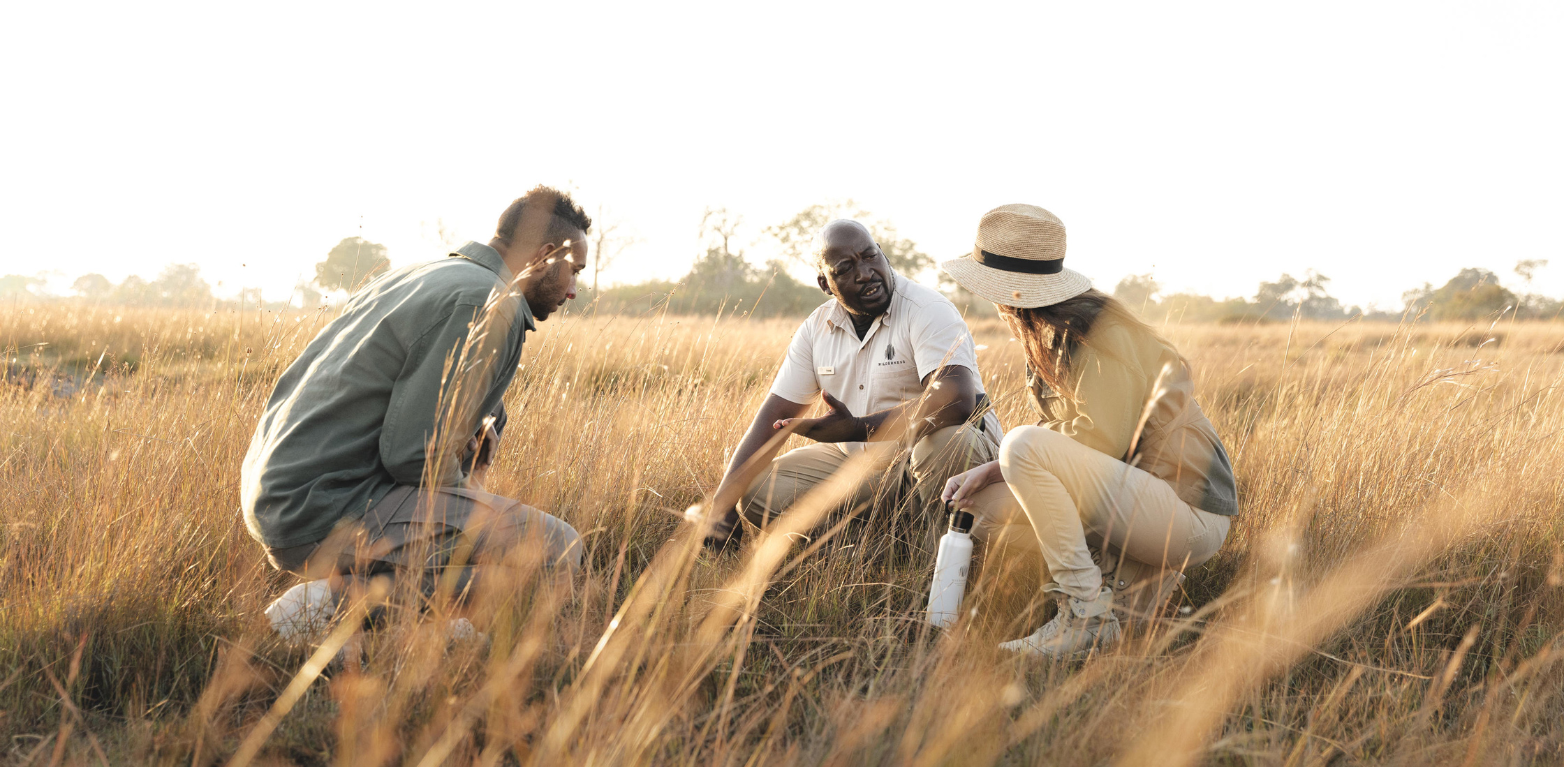 Three people kneel together through tall grass on a guided safari, with late-afternoon sun and distant trees.