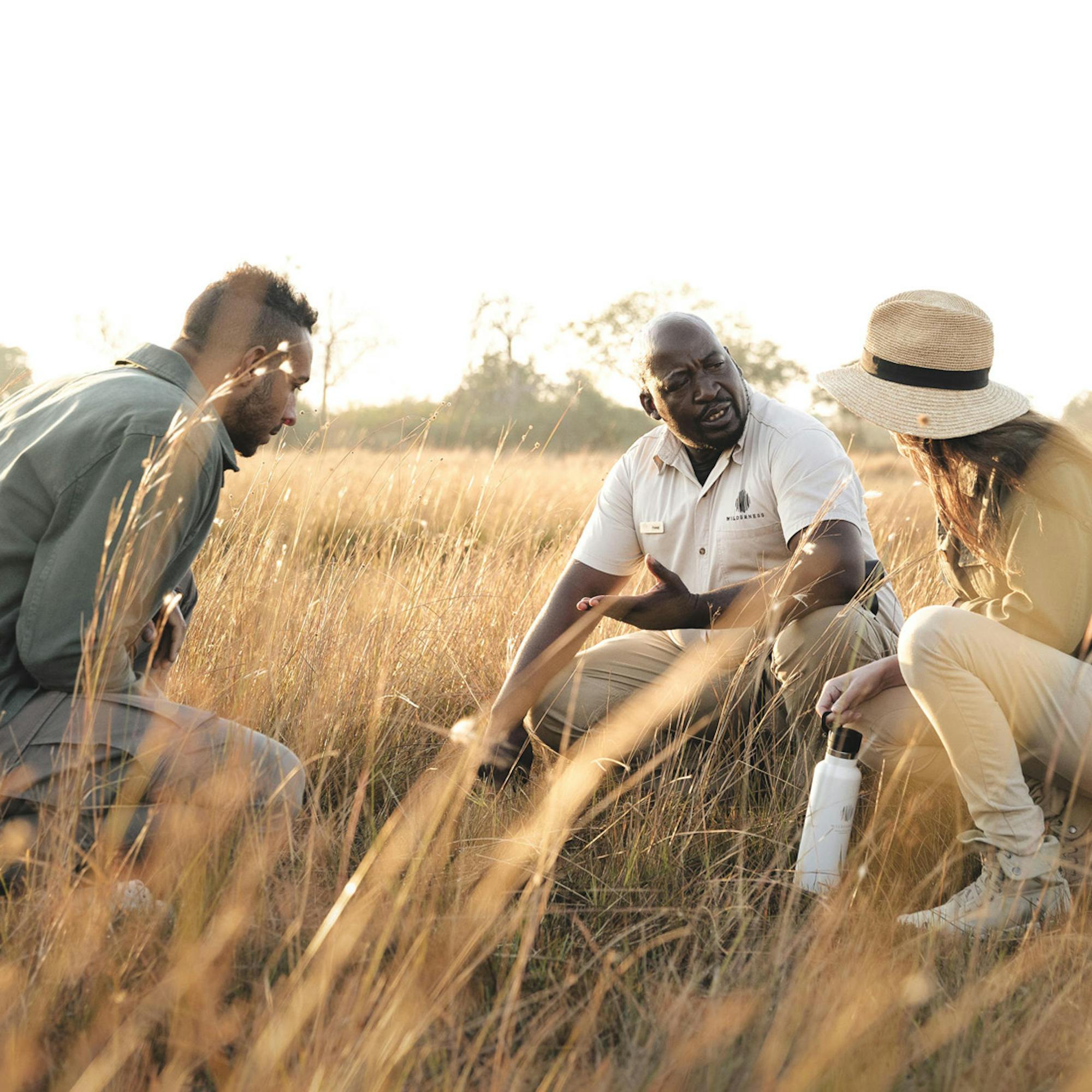 Three people kneel together through tall grass on a guided safari, with late-afternoon sun and distant trees.