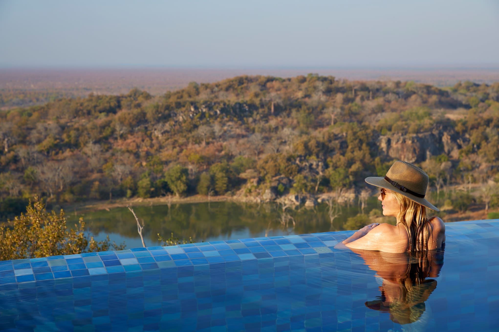 Person relaxes in an infinity pool wearing a hat, gazing over a wooded valley beyond the blue-tiled pool edge.