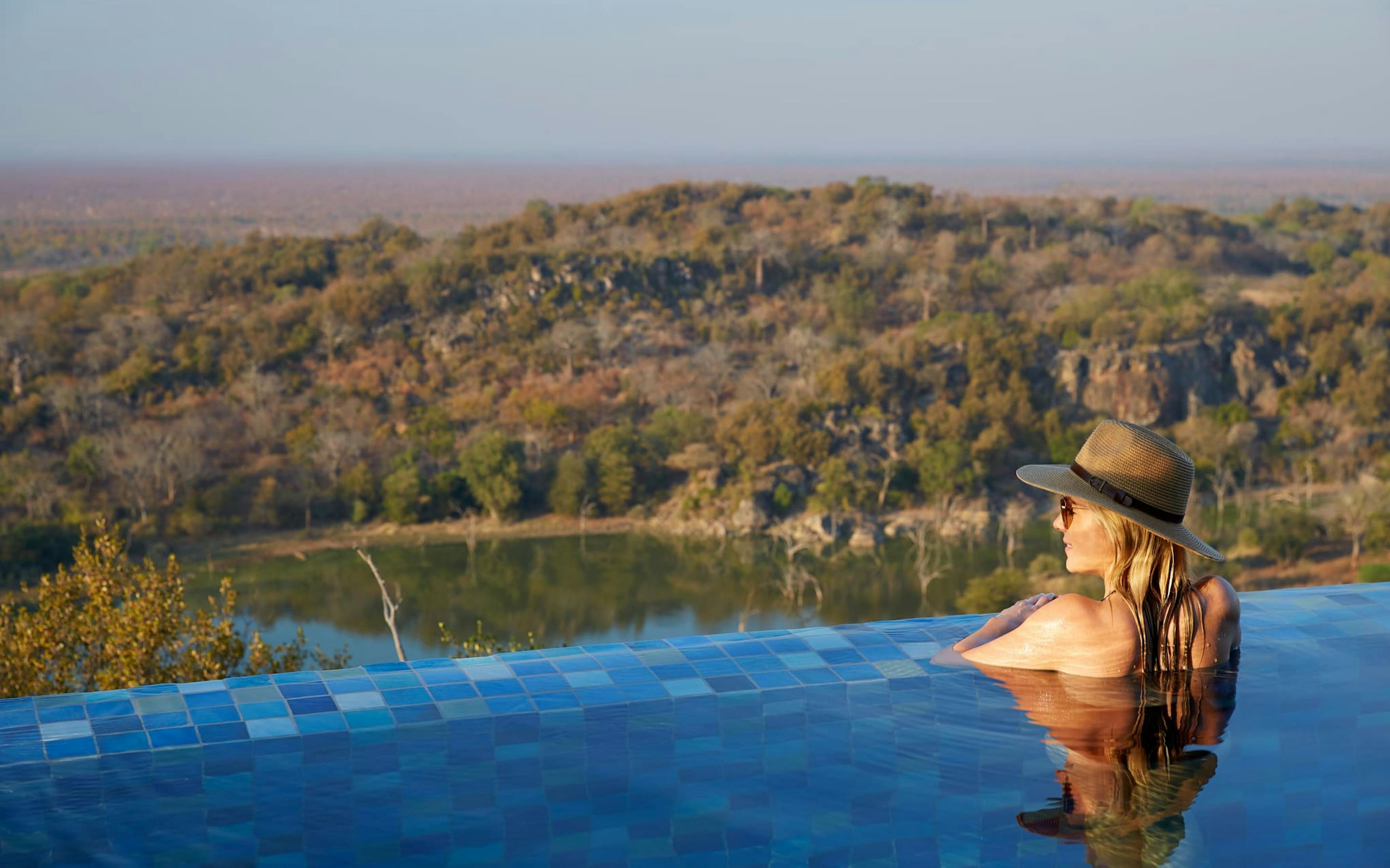 Person relaxes in an infinity pool wearing a hat, gazing over a wooded valley beyond the blue-tiled pool edge.