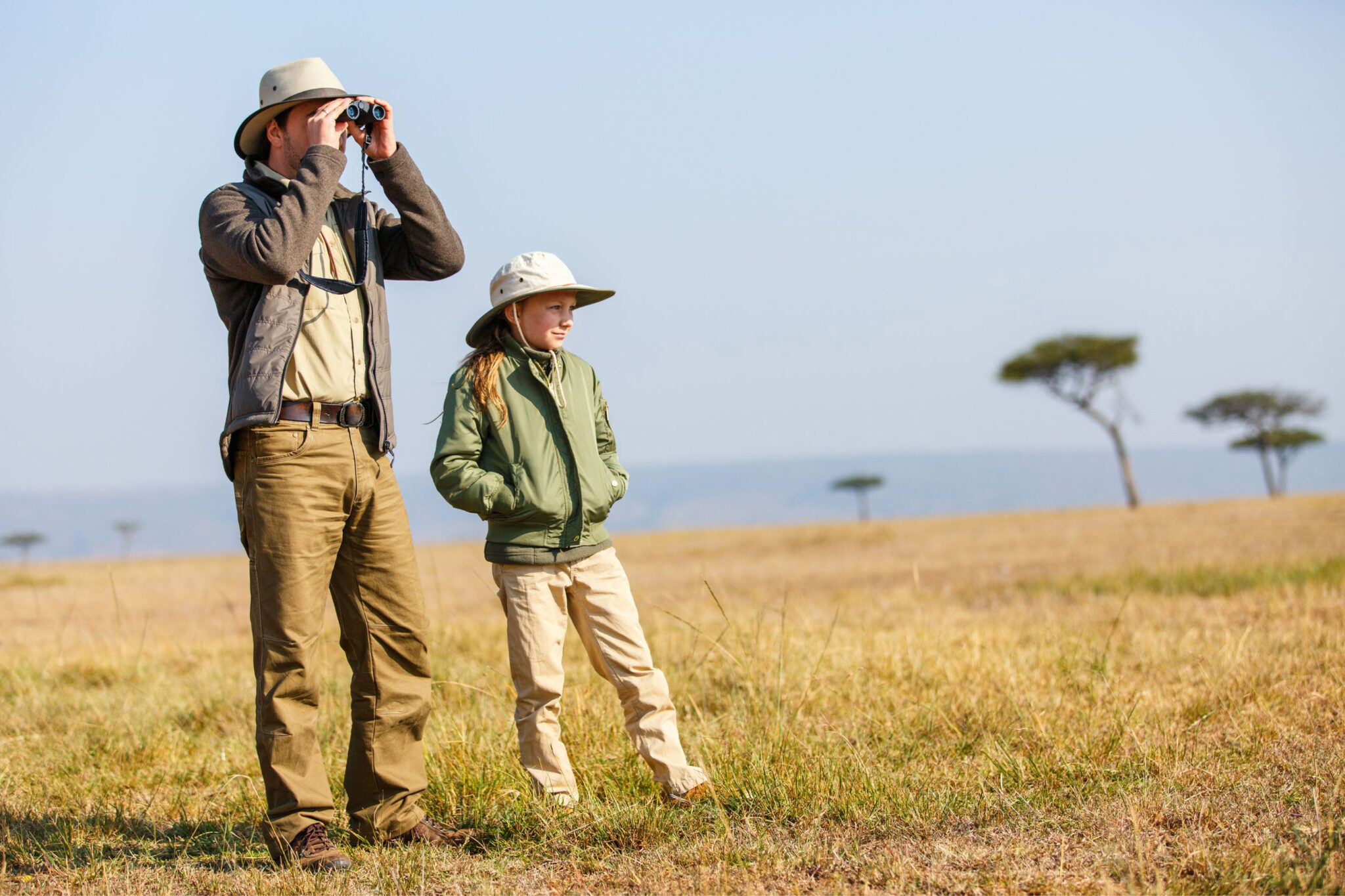Adult uses binoculars beside a child in a puffer jacket and sun hat, standing in golden savanna grass with trees ahead.