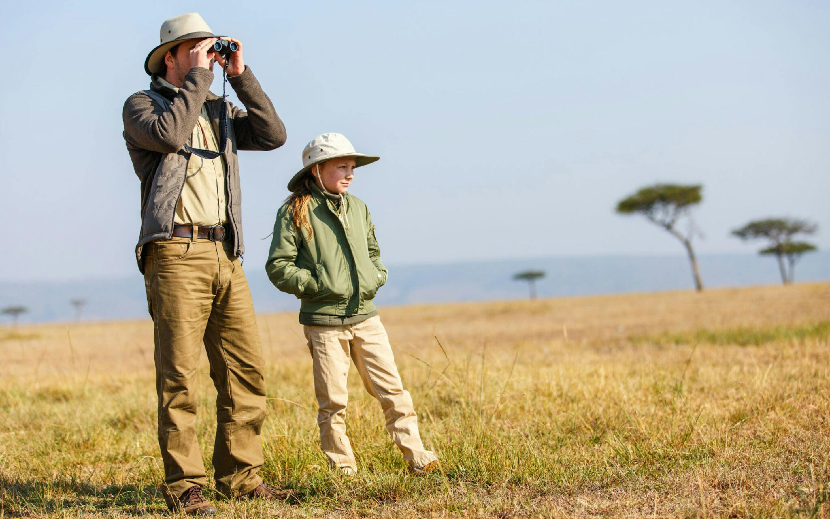 Adult uses binoculars beside a child in a puffer jacket and sun hat, standing in golden savanna grass with trees ahead.