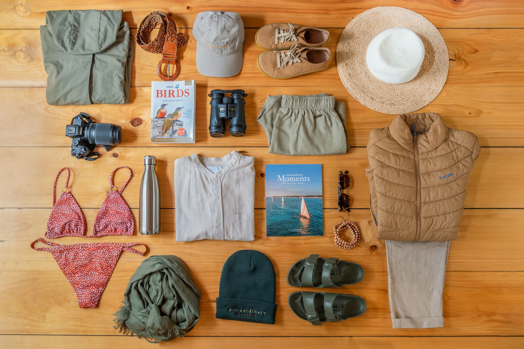 Flat lay of safari packing items on a wooden floor, neatly arranged with clothing, hat, binoculars, and toiletries.