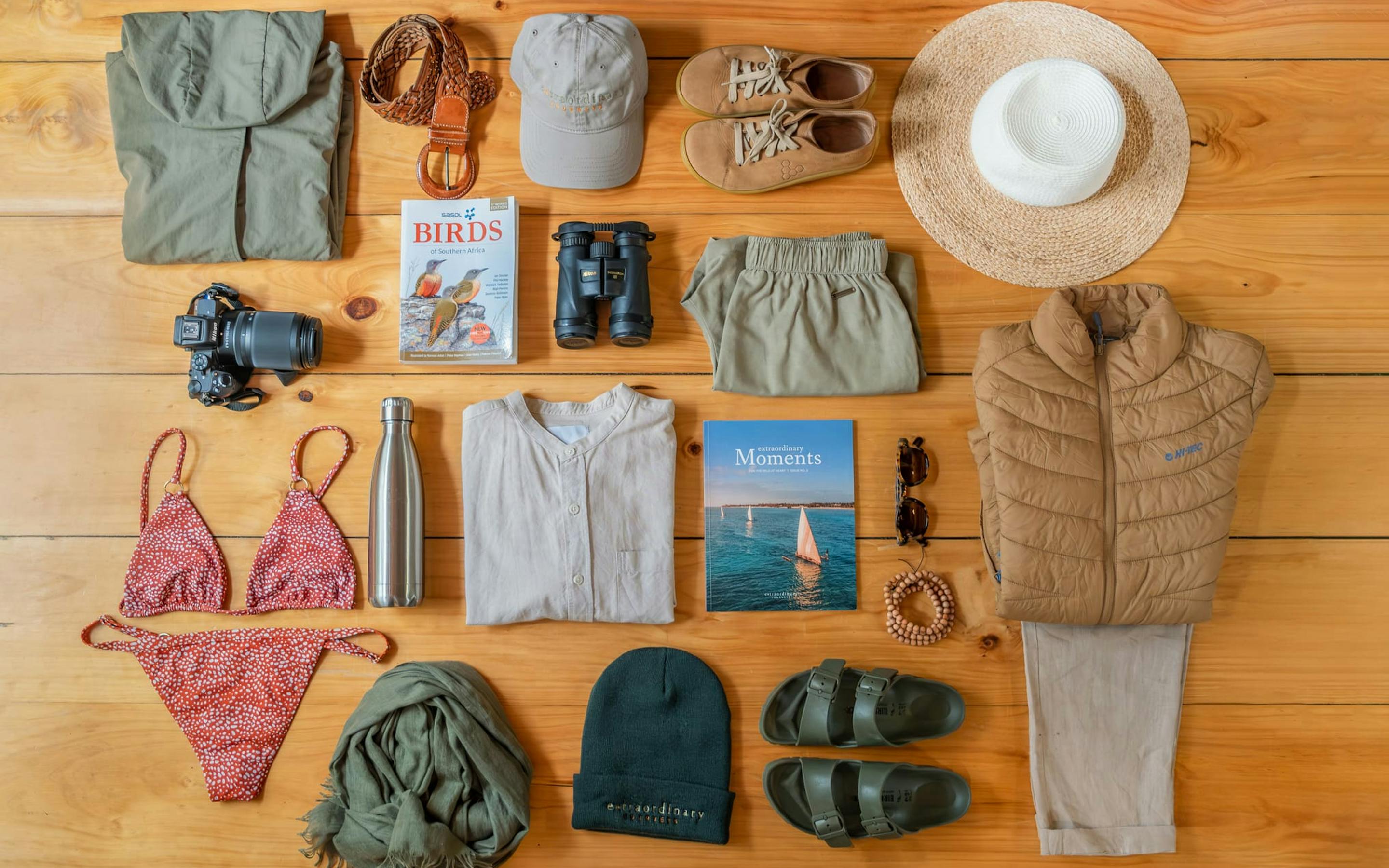 Flat lay of safari packing items on a wooden floor, neatly arranged with clothing, hat, binoculars, and toiletries.