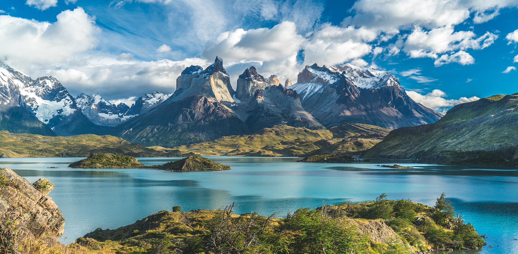Dramatic jagged peaks rise beyond a blue lake under scattered clouds, with sunlit grass in the foreground.