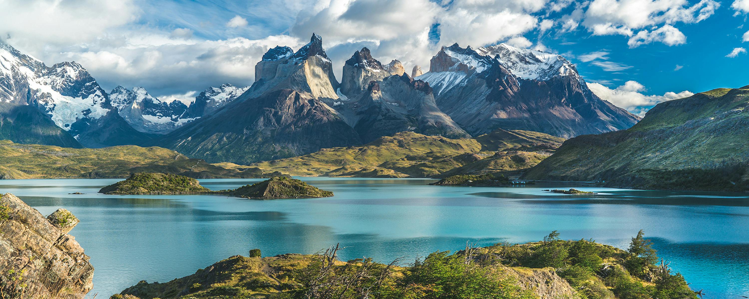 Dramatic jagged peaks rise beyond a blue lake under scattered clouds, with sunlit grass in the foreground.
