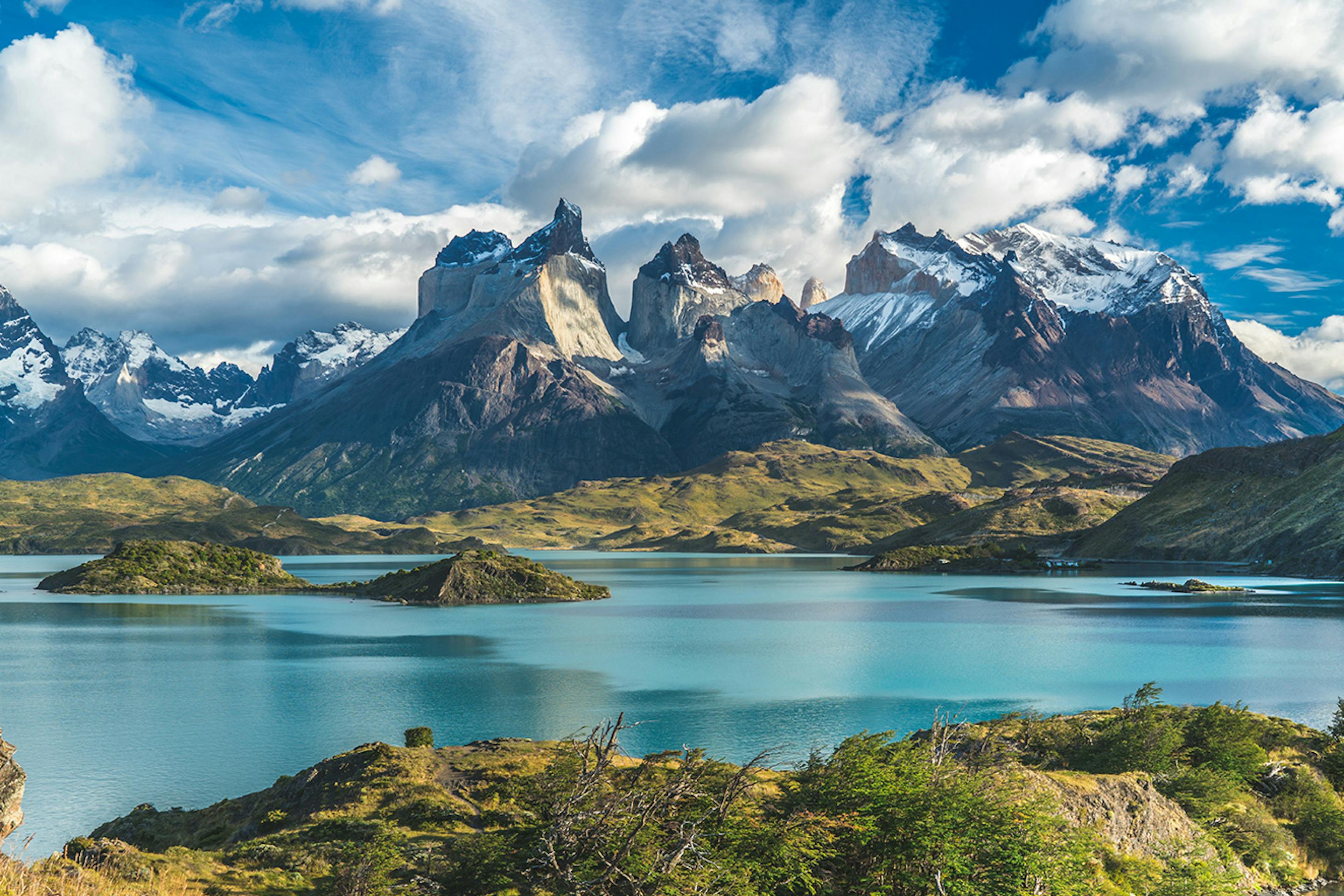 Dramatic jagged peaks rise beyond a blue lake under scattered clouds, with sunlit grass in the foreground.