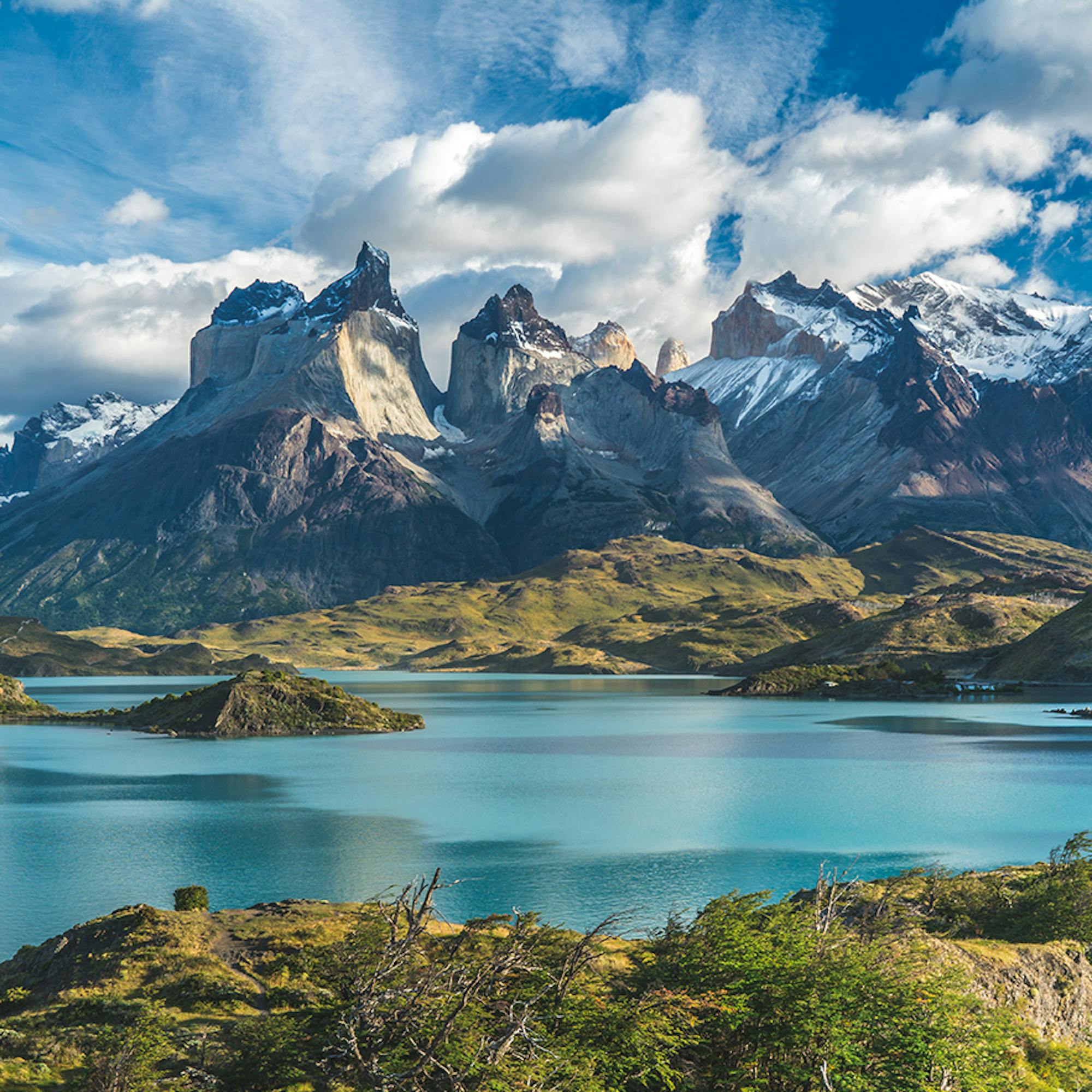 Dramatic jagged peaks rise beyond a blue lake under scattered clouds, with sunlit grass in the foreground.