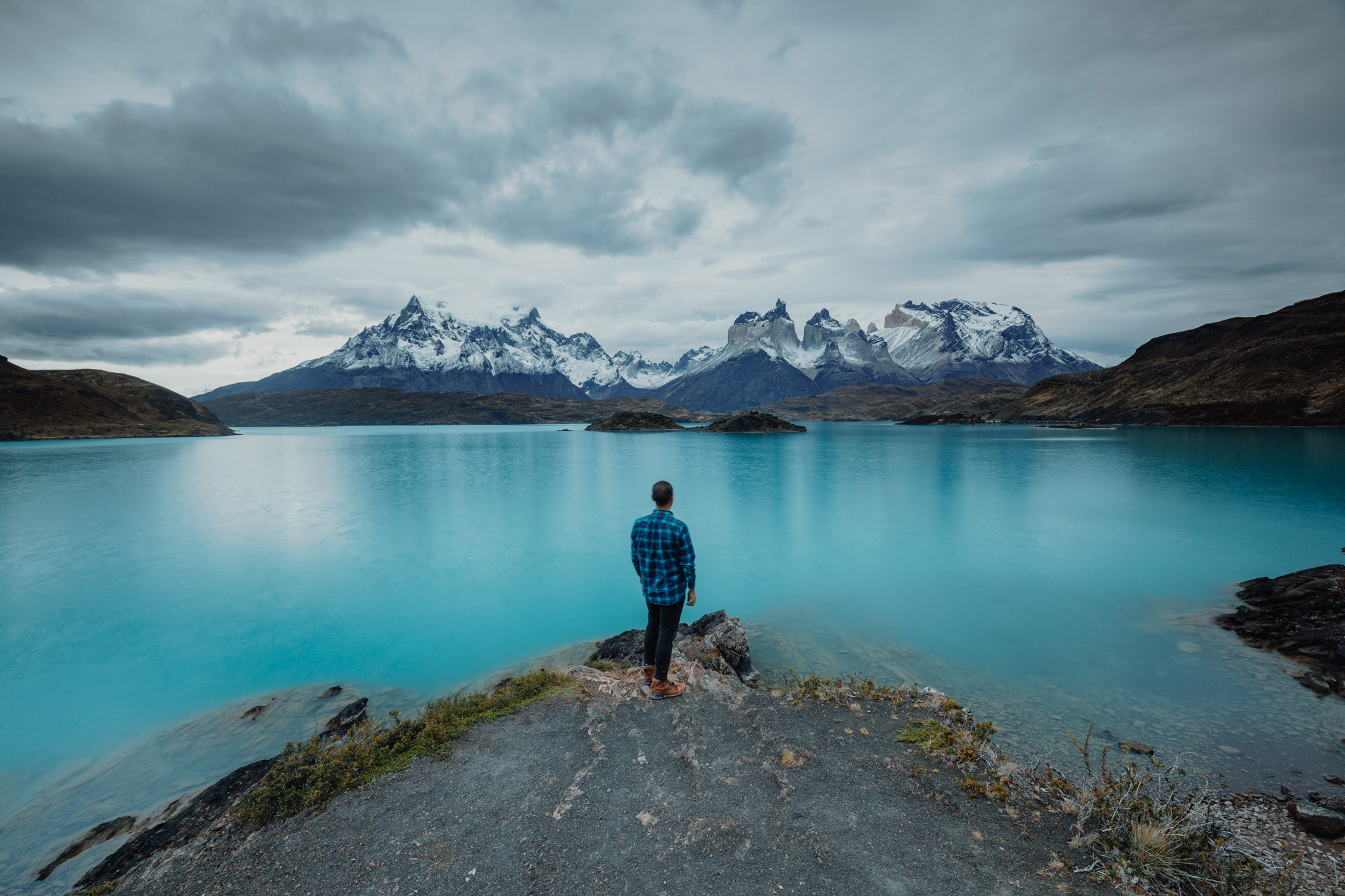 A lone hiker stands on a rocky shore facing a calm blue lake and distant snowcapped mountains under gray skies.