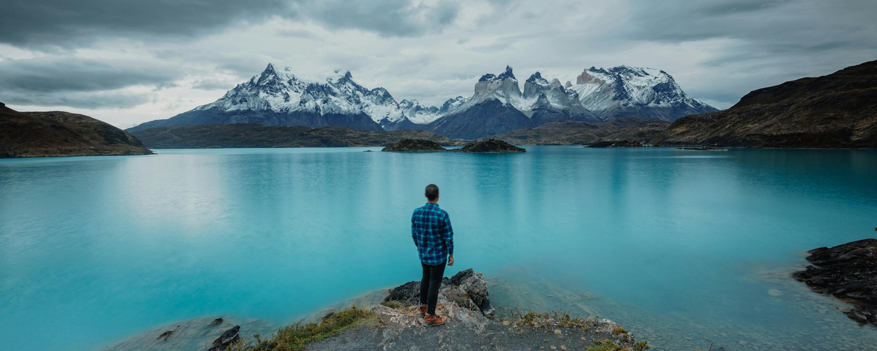 A lone hiker stands on a rocky shore facing a calm blue lake and distant snowcapped mountains under gray skies.