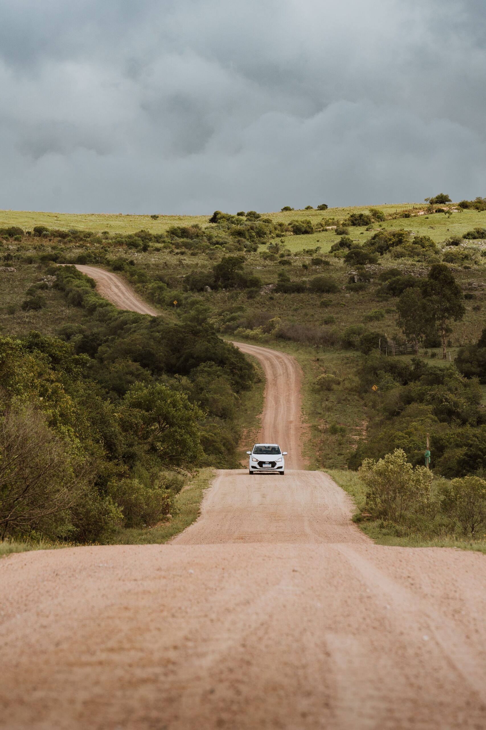 A dusty road stretches through rolling green scrub toward distant hills, with a small vehicle centered beneath gray clouds.