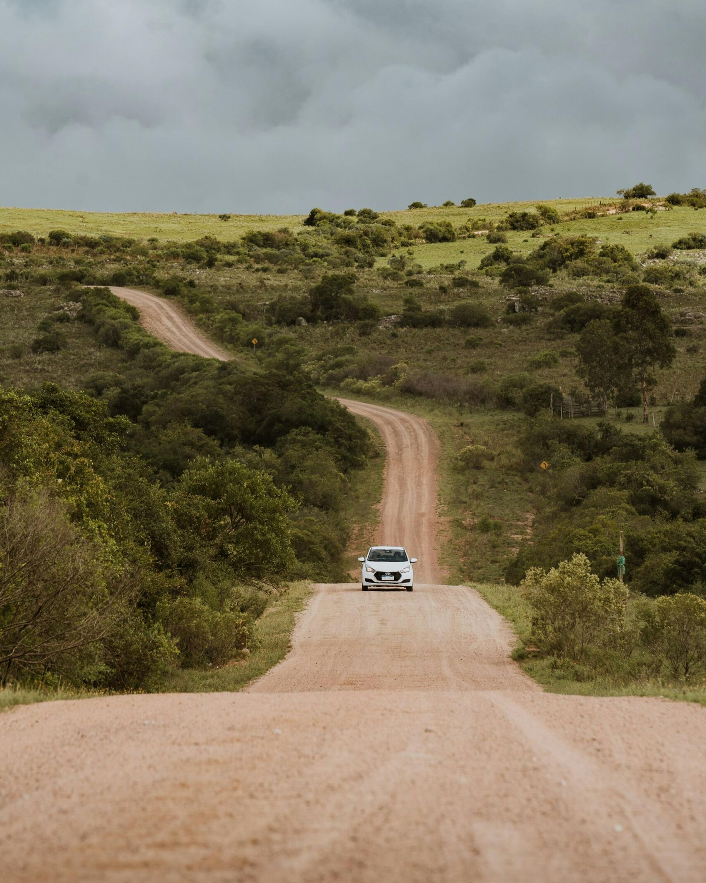 A dusty road stretches through rolling green scrub toward distant hills, with a small vehicle centered beneath gray clouds.