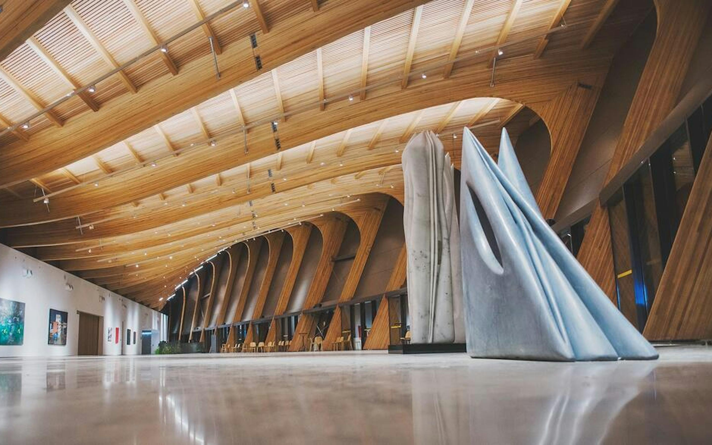 A modern gallery with curved timber ceilings surrounds a draped sculpture, with artworks spaced along bright walls.