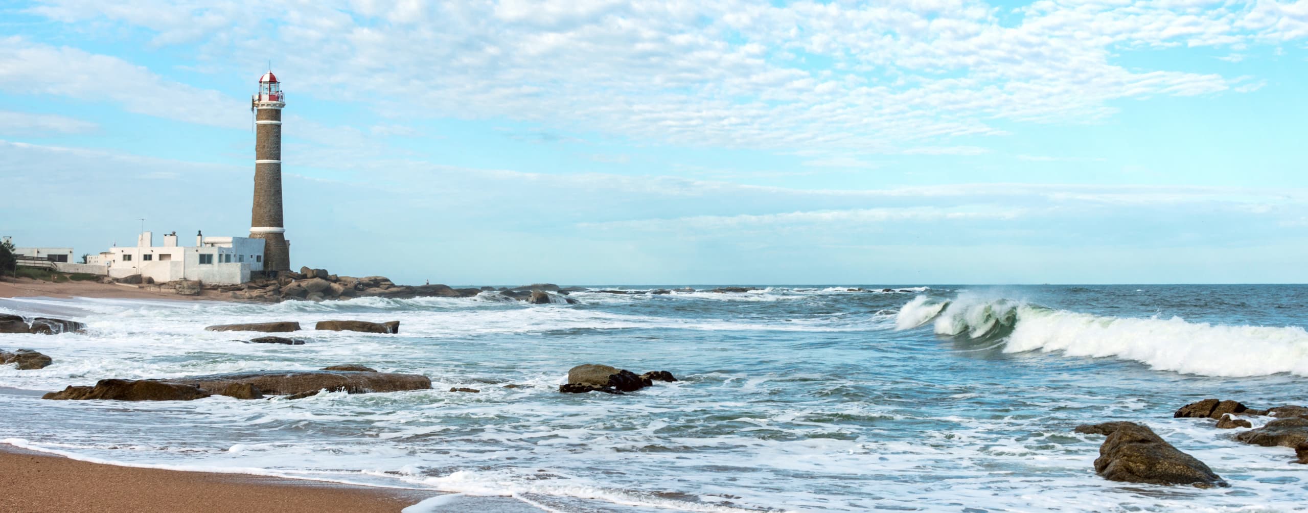 Waves break over dark shoreline rocks as a lighthouse rises in the distance beneath a pale blue sky and clouds.