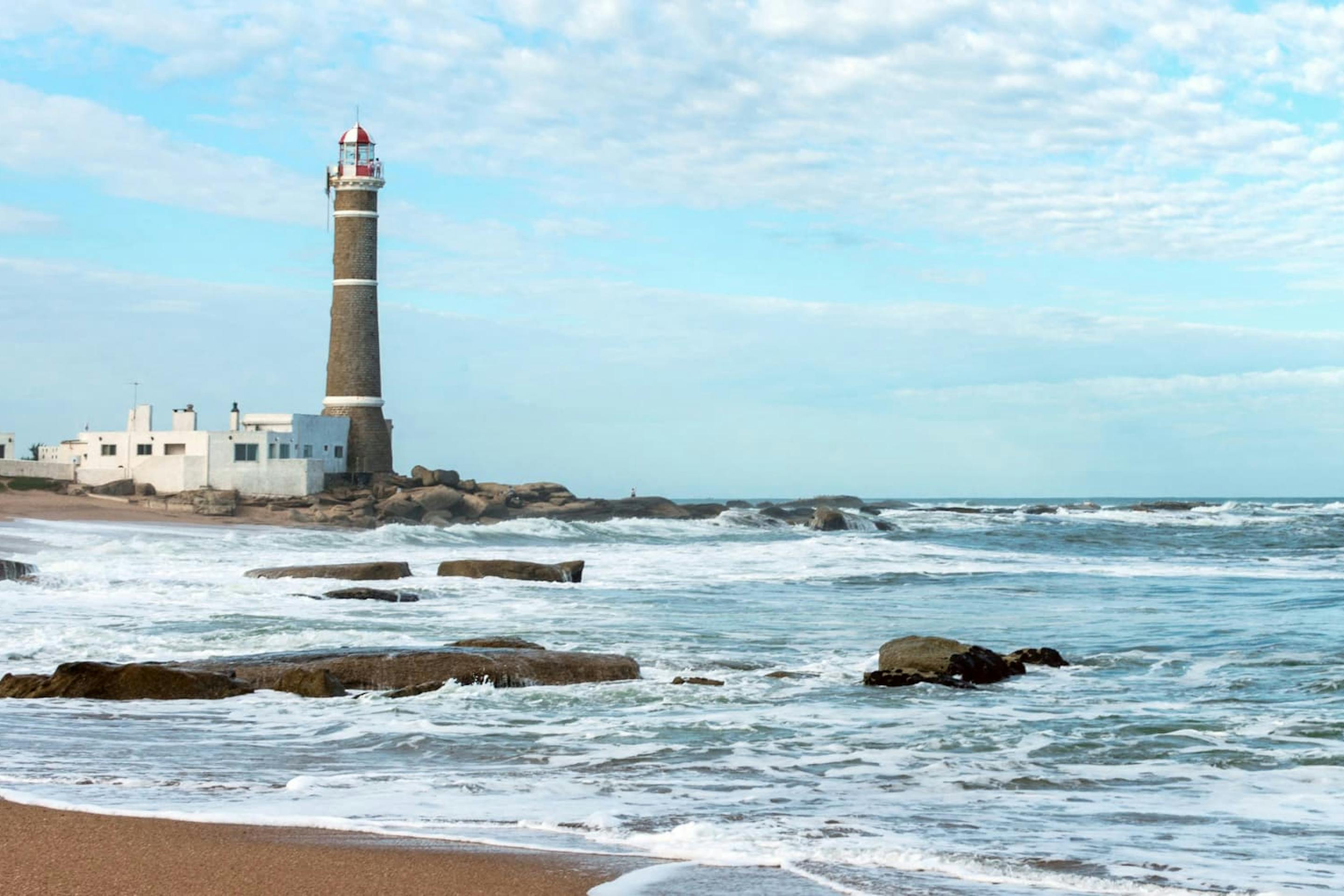 Waves break over dark shoreline rocks as a lighthouse rises in the distance beneath a pale blue sky and clouds.