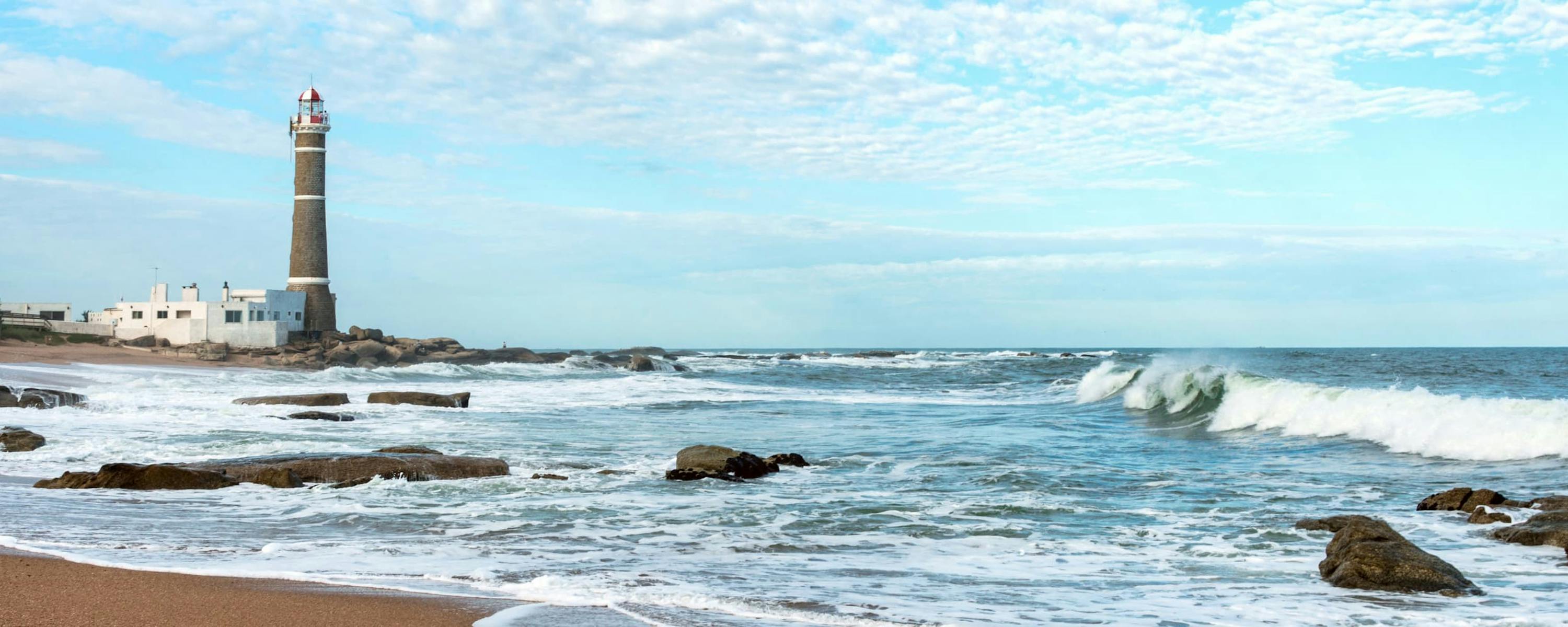 Waves break over dark shoreline rocks as a lighthouse rises in the distance beneath a pale blue sky and clouds.