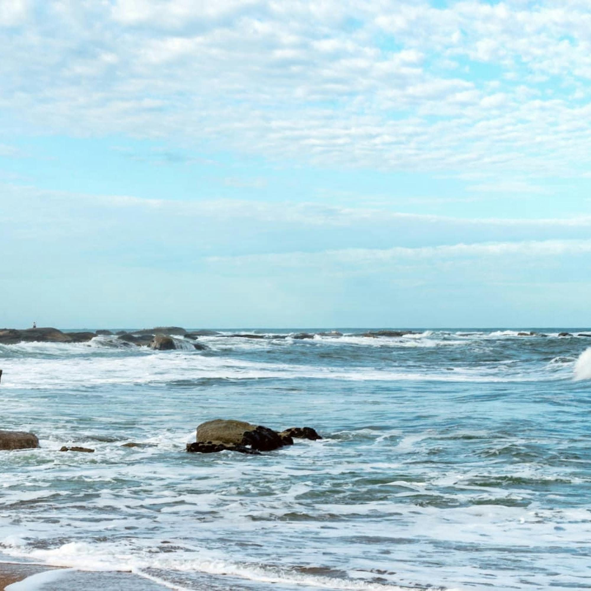 Waves break over dark shoreline rocks as a lighthouse rises in the distance beneath a pale blue sky and clouds.