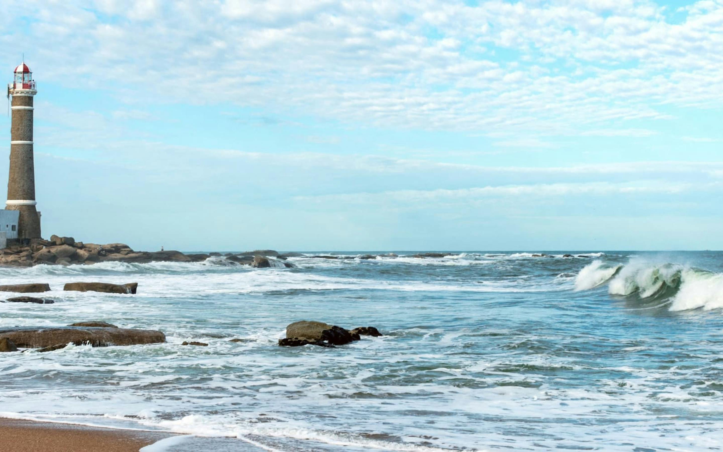 Waves break over dark shoreline rocks as a lighthouse rises in the distance beneath a pale blue sky and clouds.