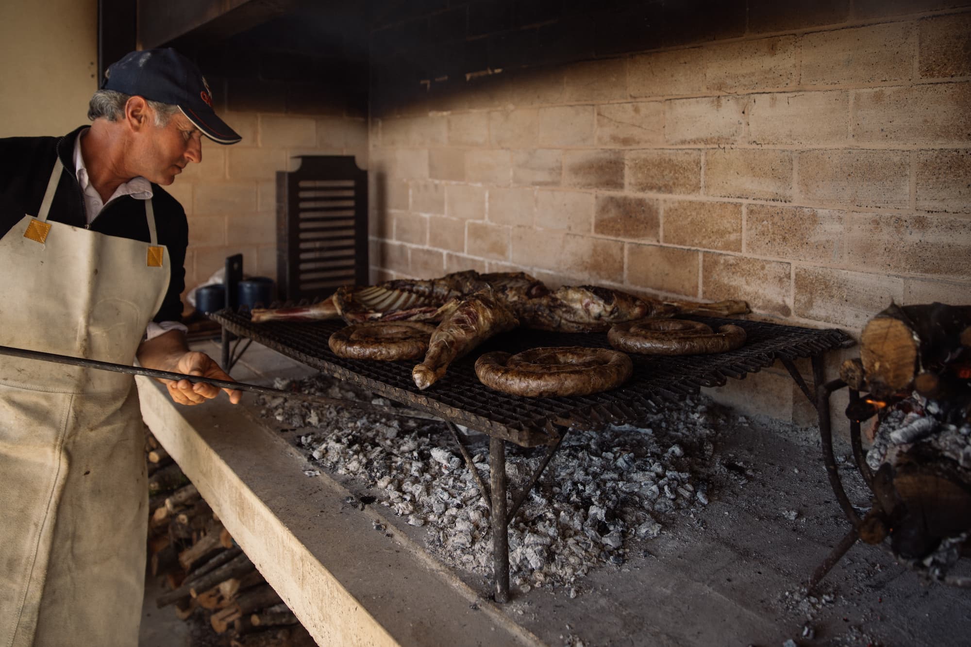 A cook grills meat over an open-fire hearth, with cast-iron pans and glowing embers inside a stone-walled kitchen.