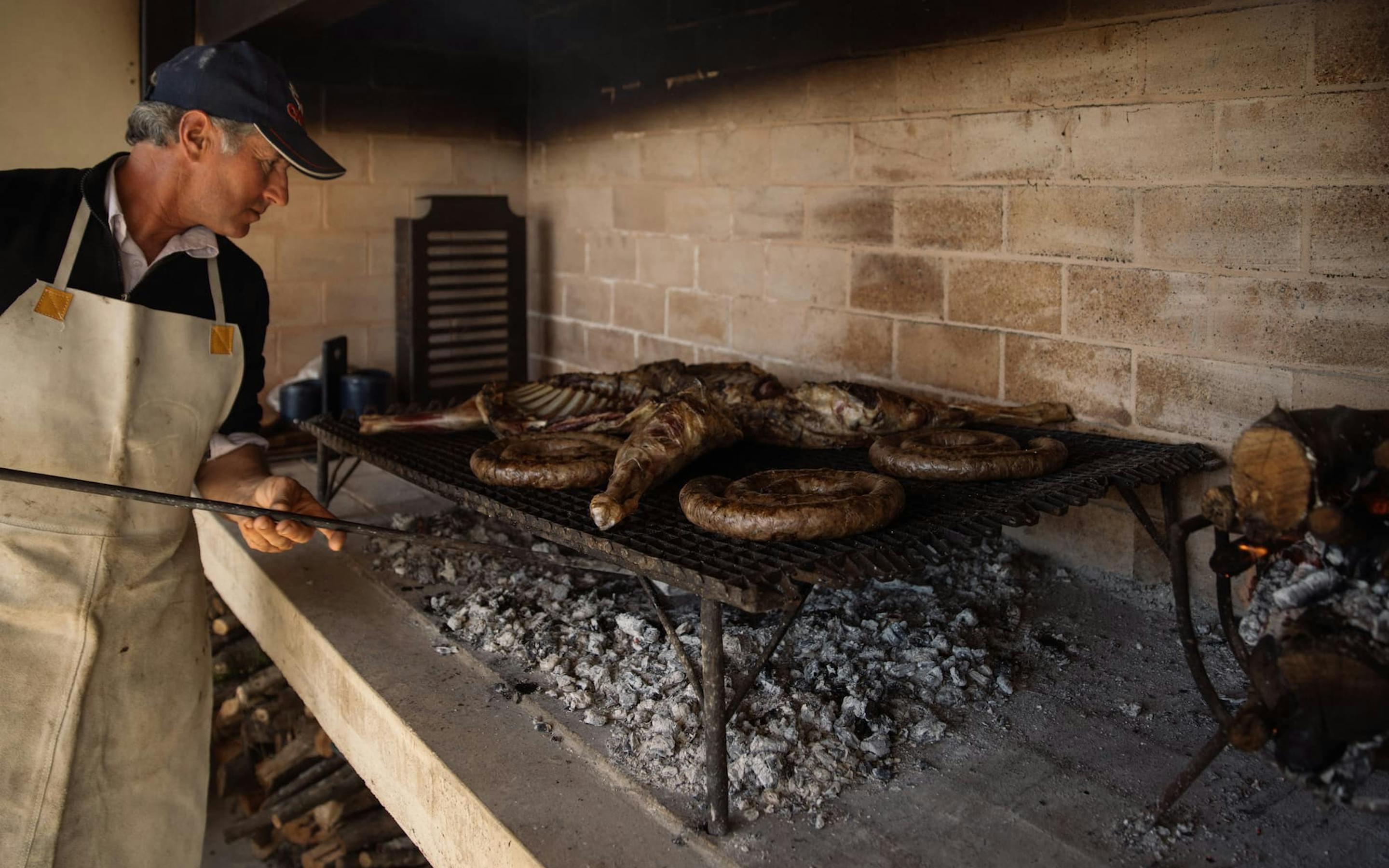 A cook grills meat over an open-fire hearth, with cast-iron pans and glowing embers inside a stone-walled kitchen.