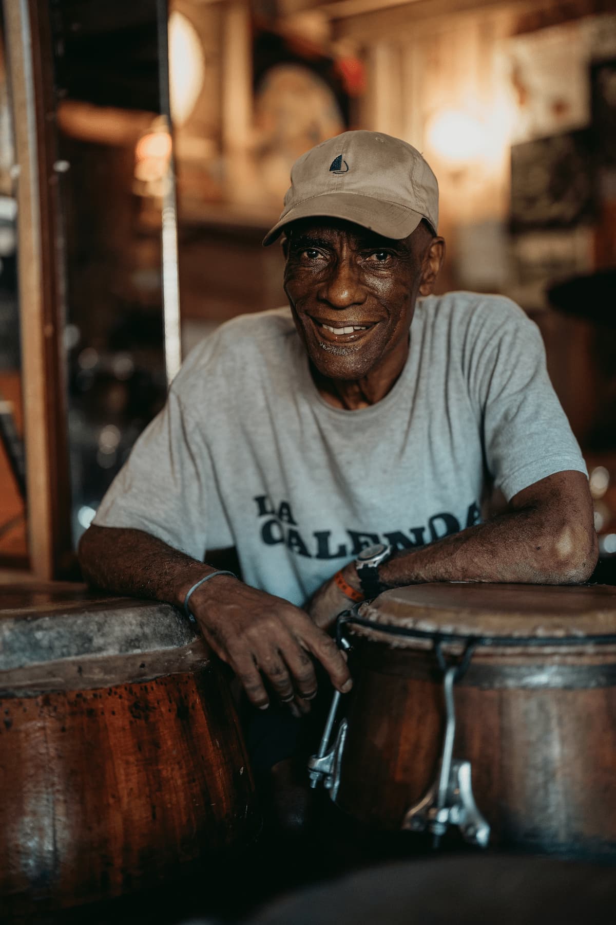 A seated drummer smiles in a dim bar beside hand drums, with warm lights reflecting off dark wood and glass.