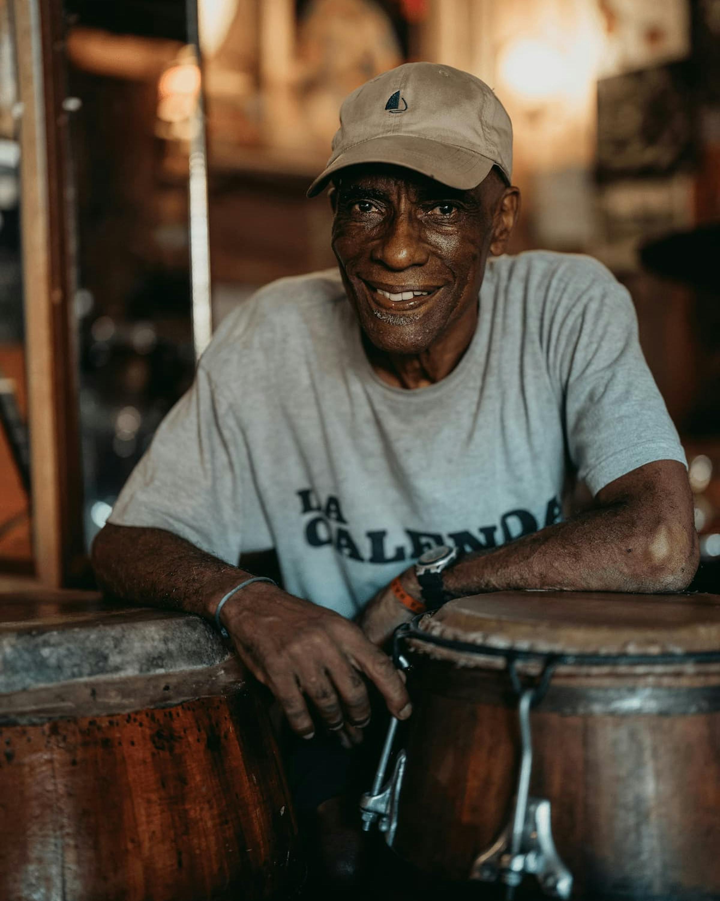 A seated drummer smiles in a dim bar beside hand drums, with warm lights reflecting off dark wood and glass.