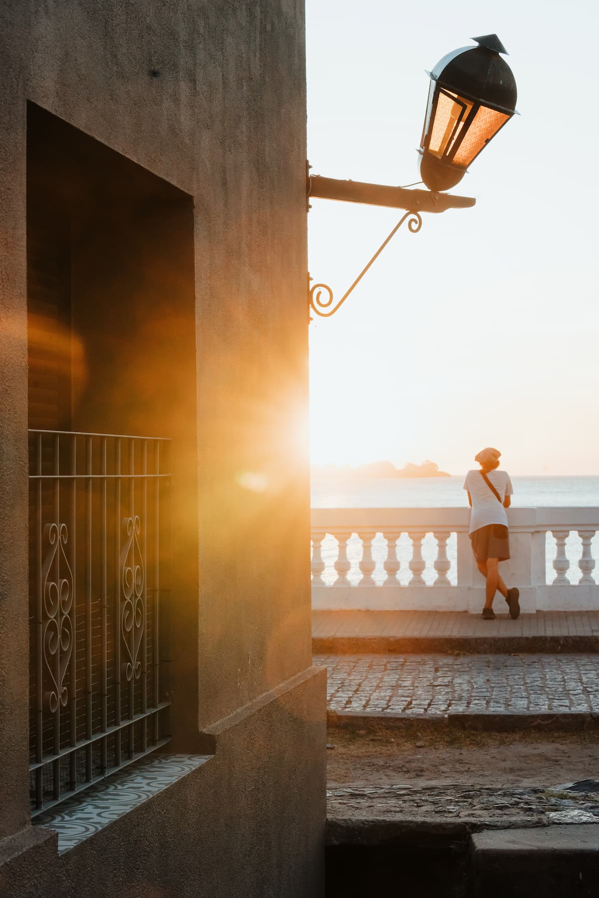 Warm sunset light spills down a narrow walkway beside the sea, with a lamppost and wooden rail silhouetted.
