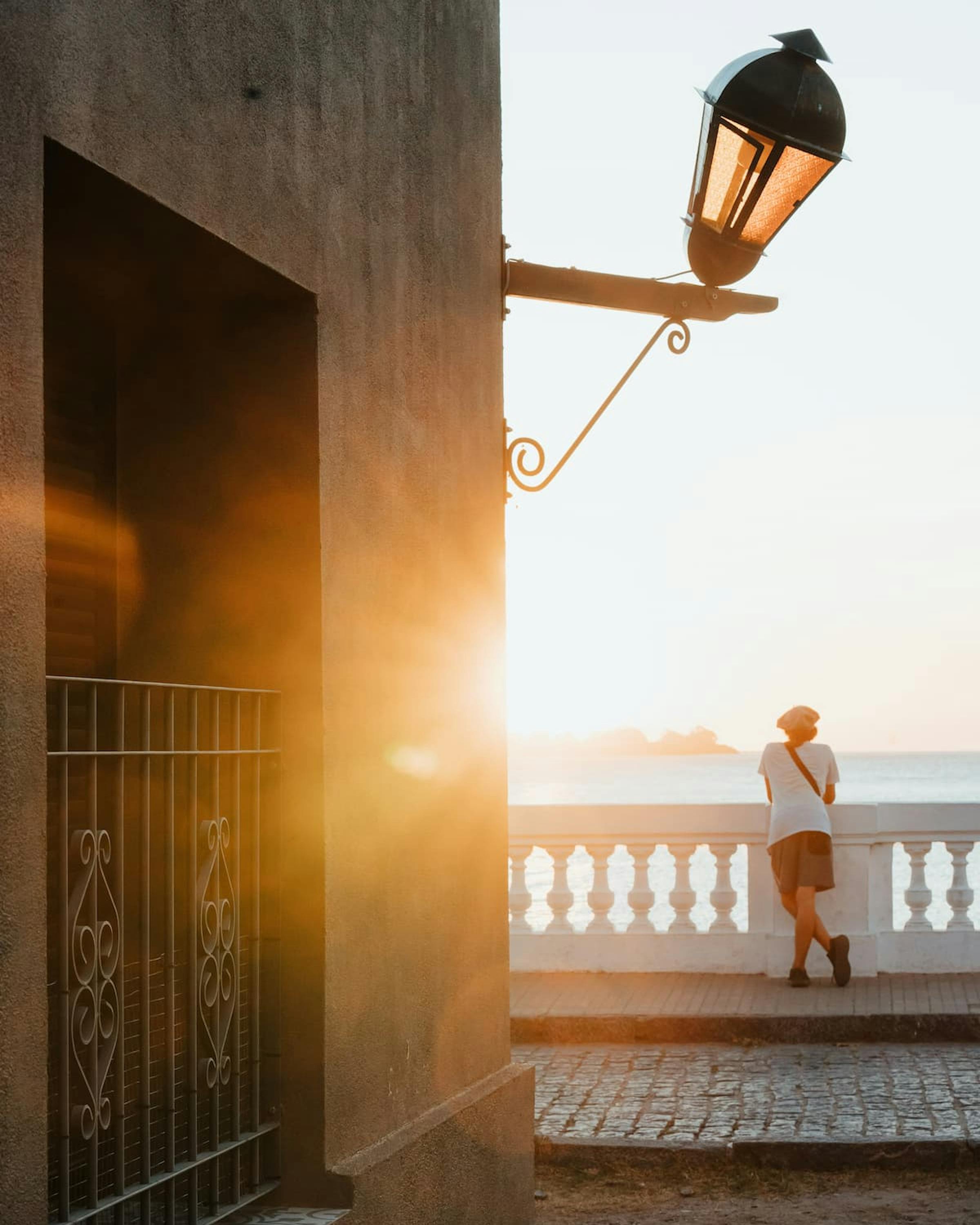Warm sunset light spills down a narrow walkway beside the sea, with a lamppost and wooden rail silhouetted.