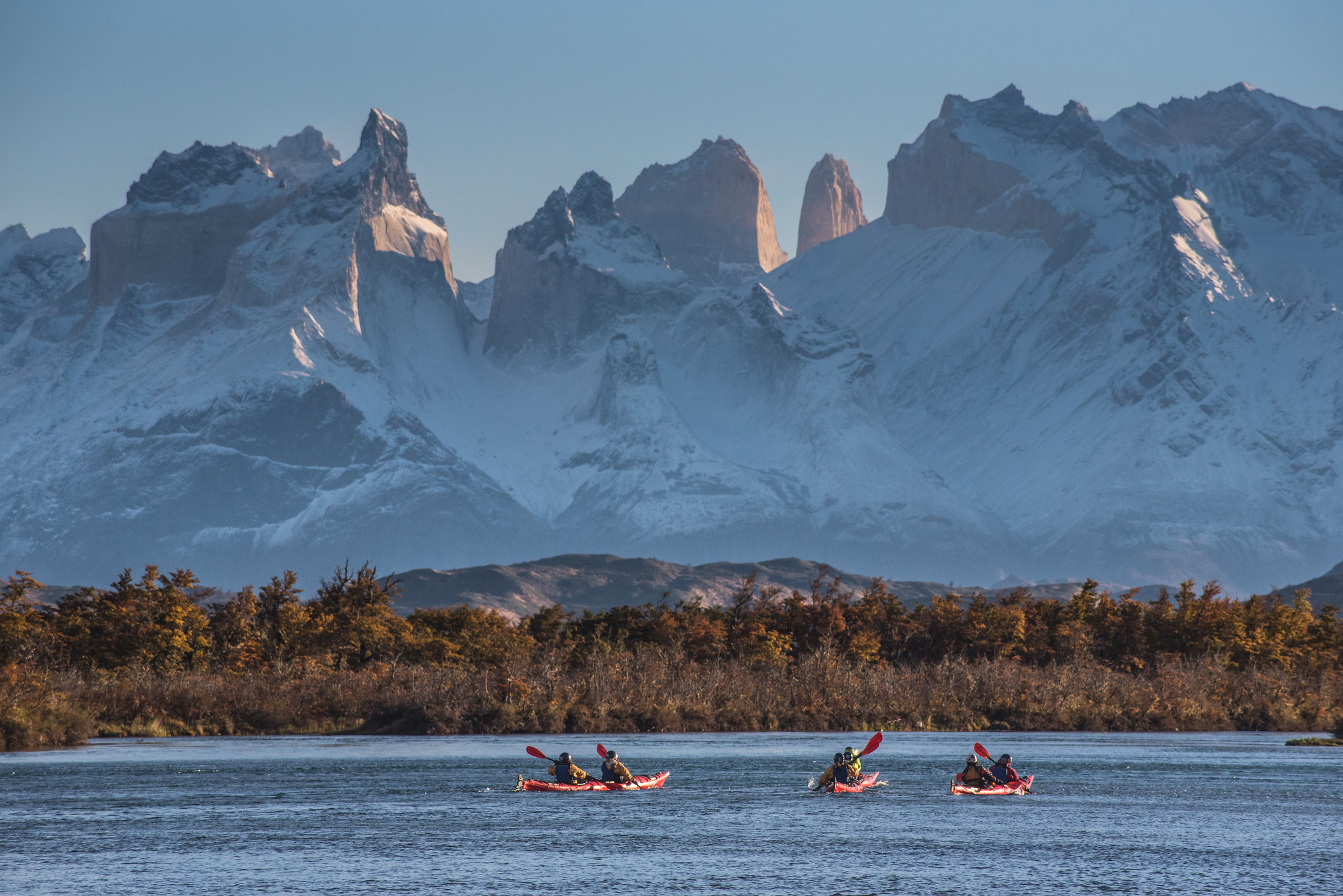 Kayakers paddle a wide lake, with snowy mountains and the Torres del Paine skyline beyond dark forest.