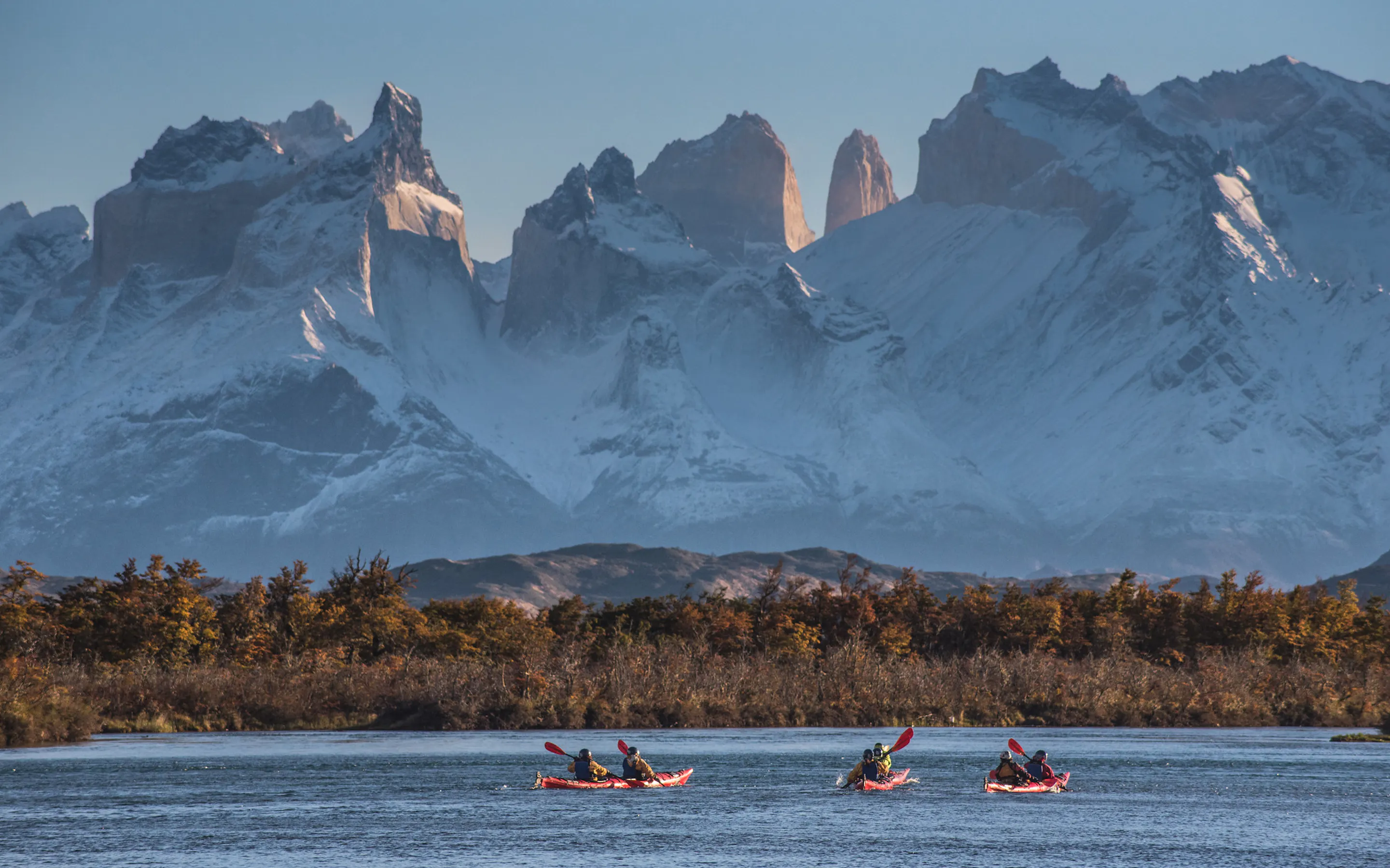Kayakers paddle a wide lake, with snowy mountains and the Torres del Paine skyline beyond dark forest.