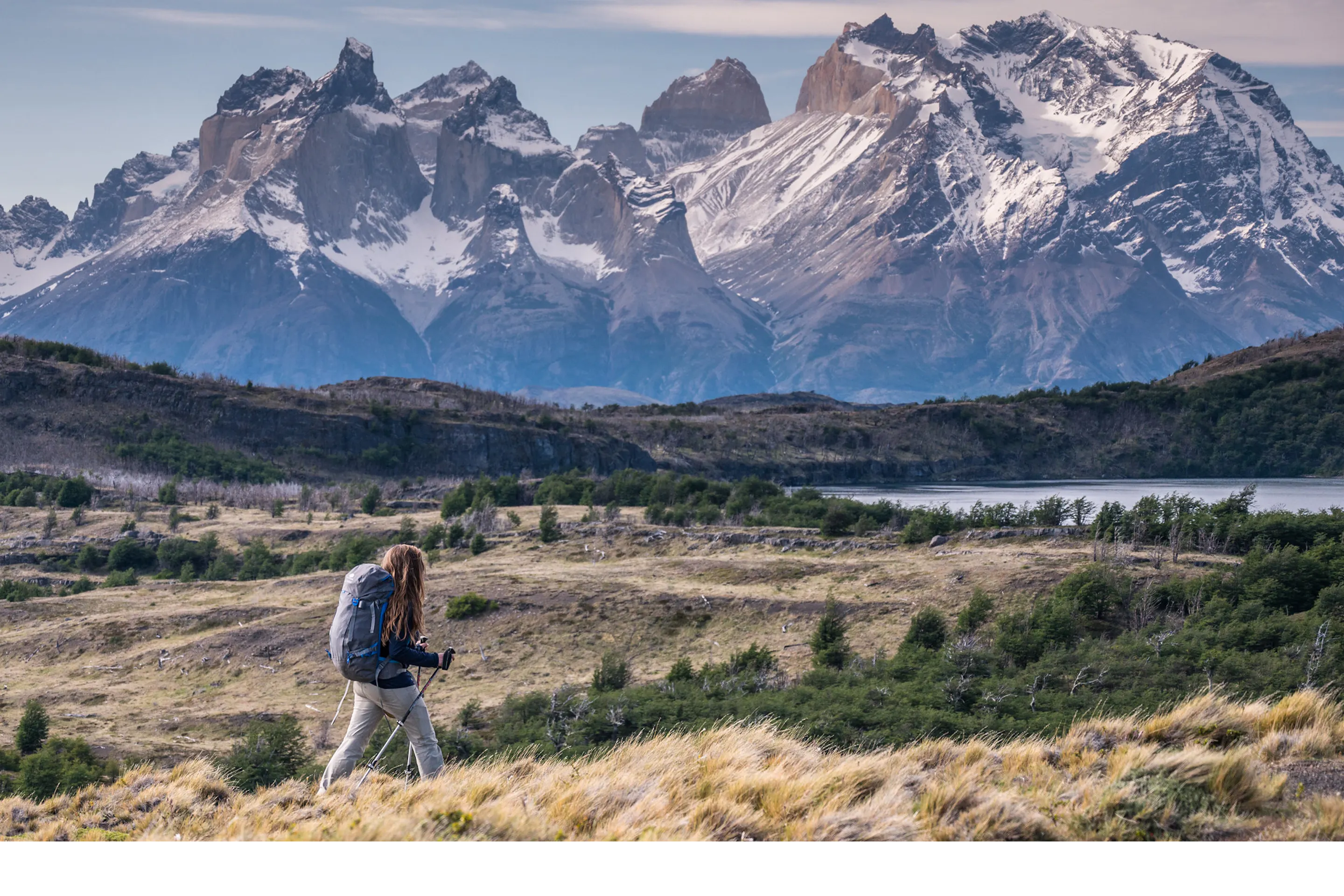 A hiker crosses open grassland with a backpack, jagged Torres del Paine peaks rising beyond the steppe, in Patagonia..