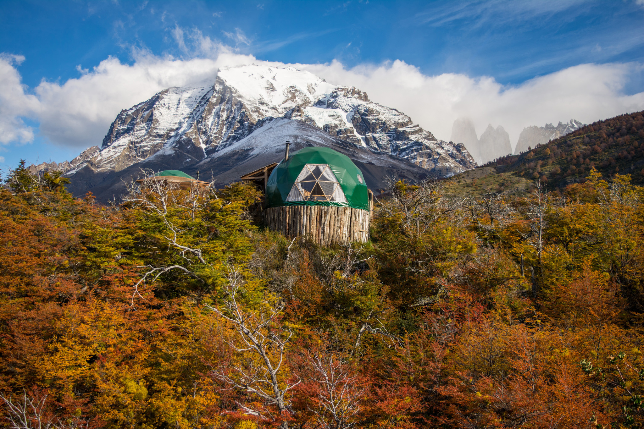 A green suite dome sits above autumn trees, backed by a snowcapped peak and clear Patagonian sky, with rugged peaks beyond.